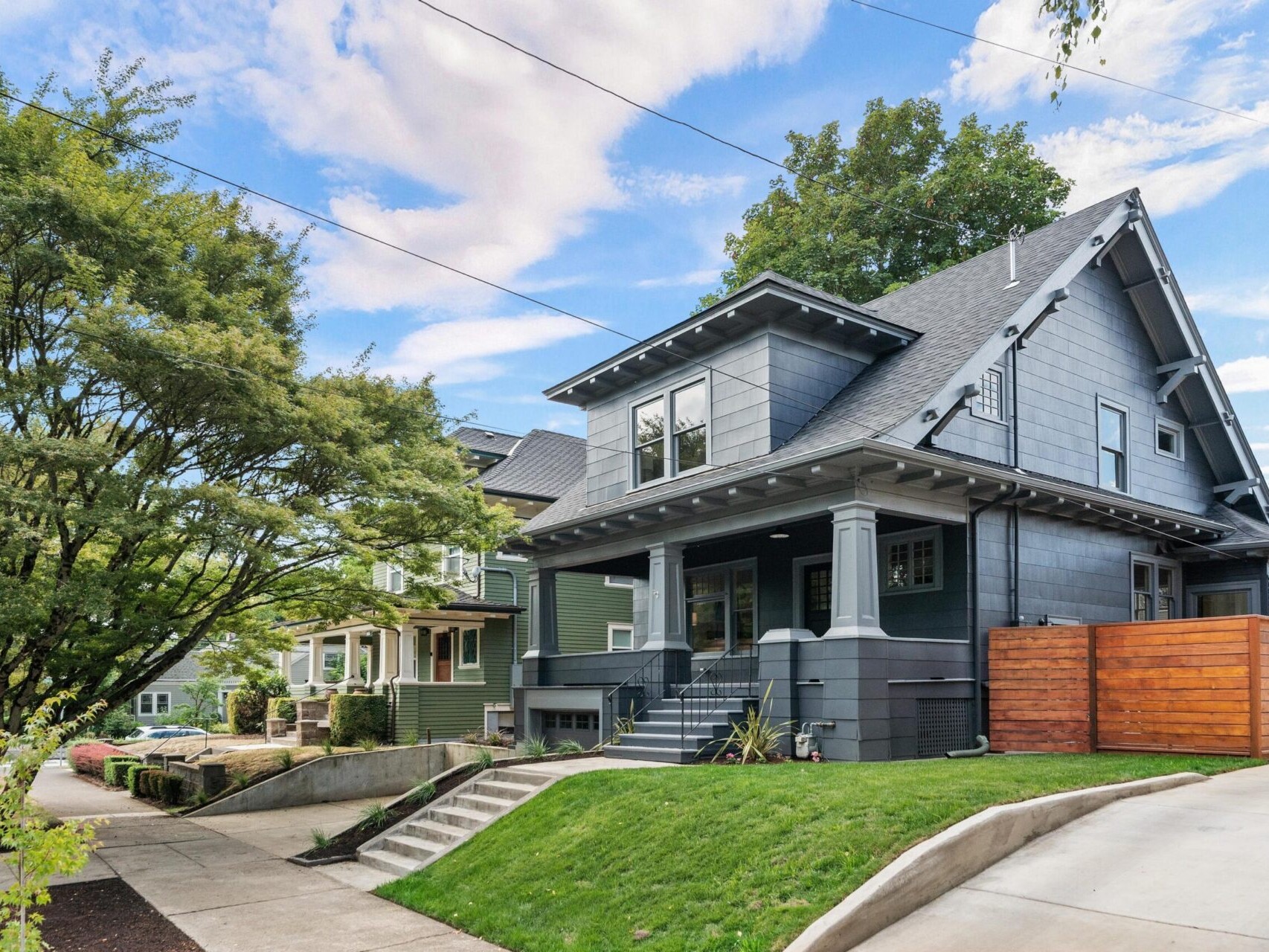A modern gray house with a gabled roof, white trim, and a covered front porch graces this Portland, Oregon neighborhood. Nestled in a neatly landscaped yard with a wooden fence to the right and a towering tree on the left, neighboring houses peek out under the expansive blue sky.