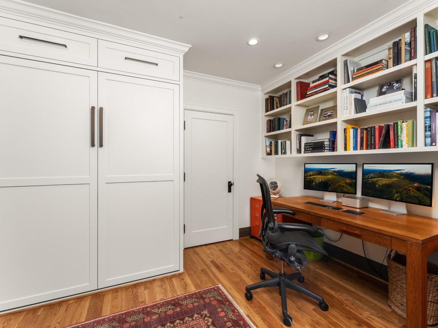 A home office with wooden flooring, featuring a large desk with dual monitors and a swivel chair. Built-in shelves are filled with books and decor. A closed Murphy bed is on the left, and a patterned rug is on the floor—a true gem for any Portland Oregon real estate enthusiast.
