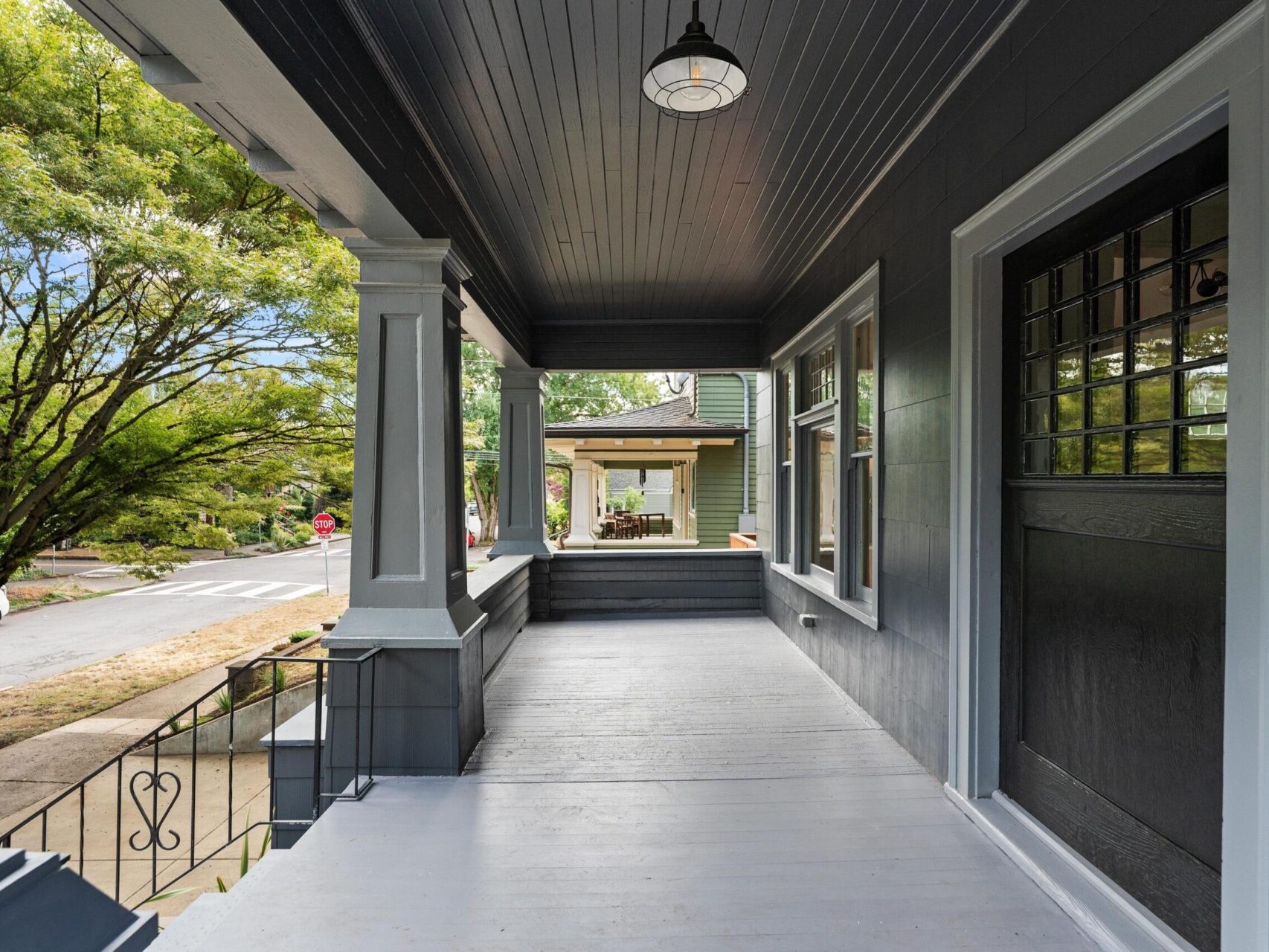 A covered porch with a dark ceiling and light-colored floor, featuring columns and a railing, overlooks a quiet Portland, Oregon street lined with trees. Adjacent to a green house, a hanging light fixture adds charm to this serene space.
