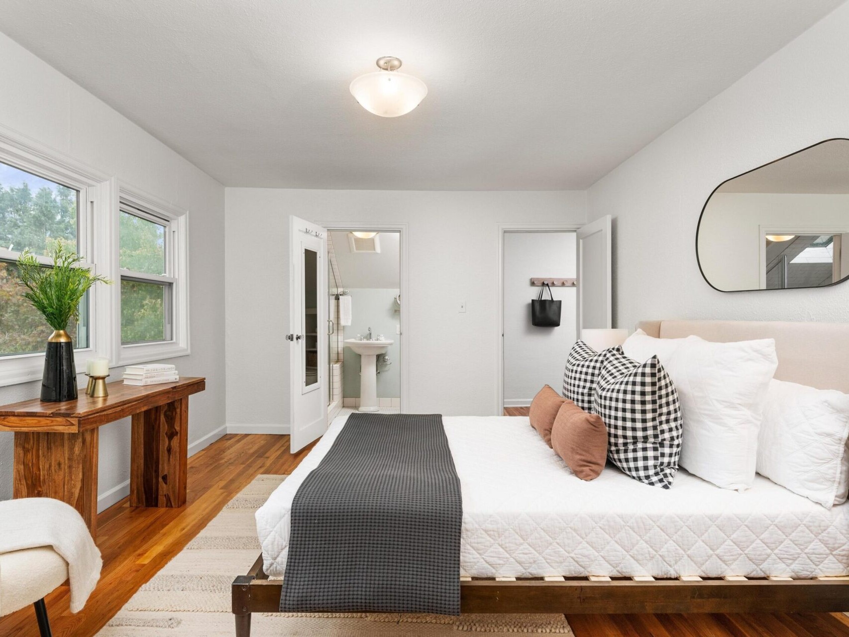 A modern bedroom with a neatly made bed featuring pillows in white and plaid. A large mirror hangs on the wall above the bed. A wooden desk with decor sits by the window, and the view leads to a bathroom through an open door. Neutral tones dominate the room.