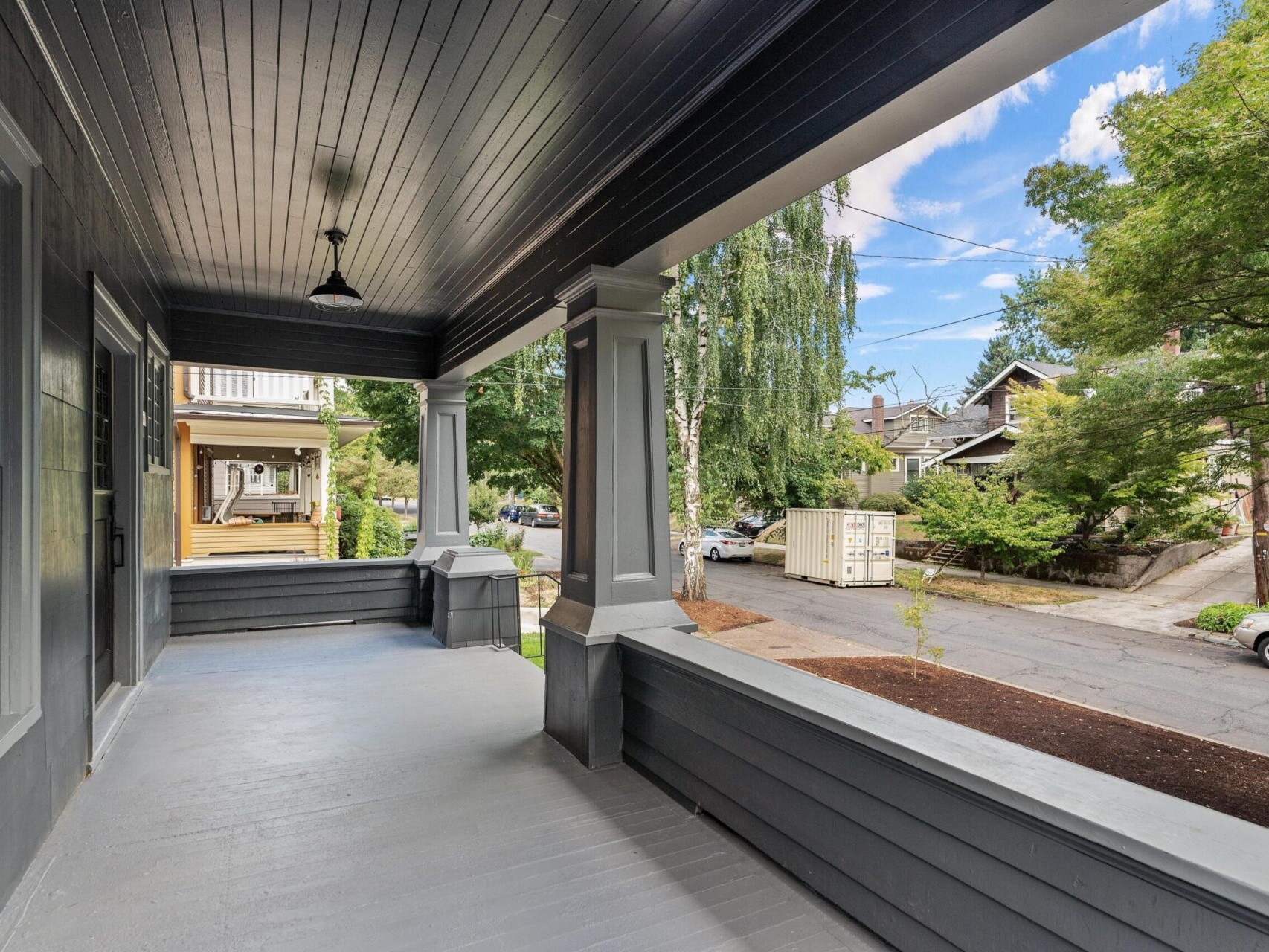 A spacious, covered front porch with gray wooden flooring and railings, overlooking a charming Portland, Oregon street lined with trees and houses. The porch features large columns and a hanging light fixture, offering a tranquil and inviting outdoor space.