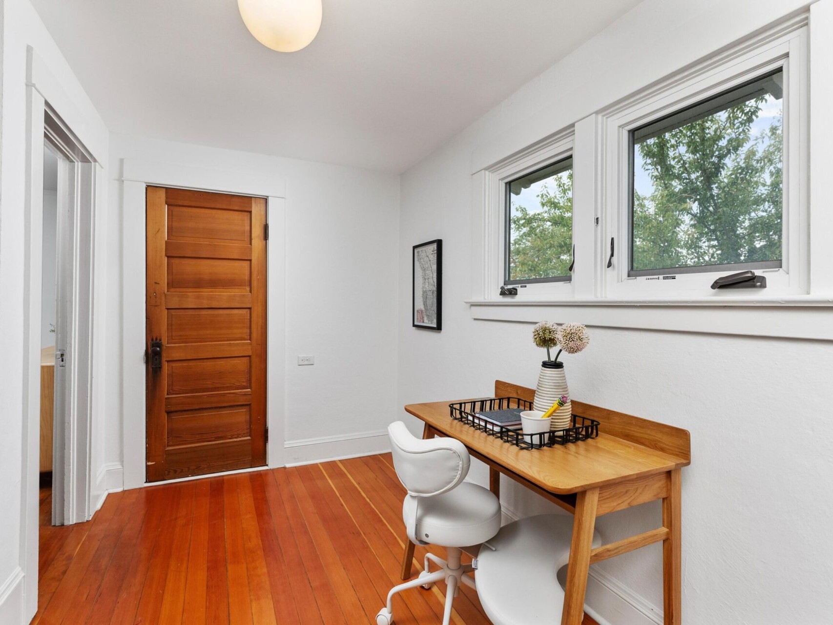 A small study area with a wooden desk and white chair is nestled next to two windows showcasing Portland Oregon's lush greenery. The room boasts hardwood floors, a wooden door, and pristine white walls. A round ceiling light beautifully illuminates the inviting space.