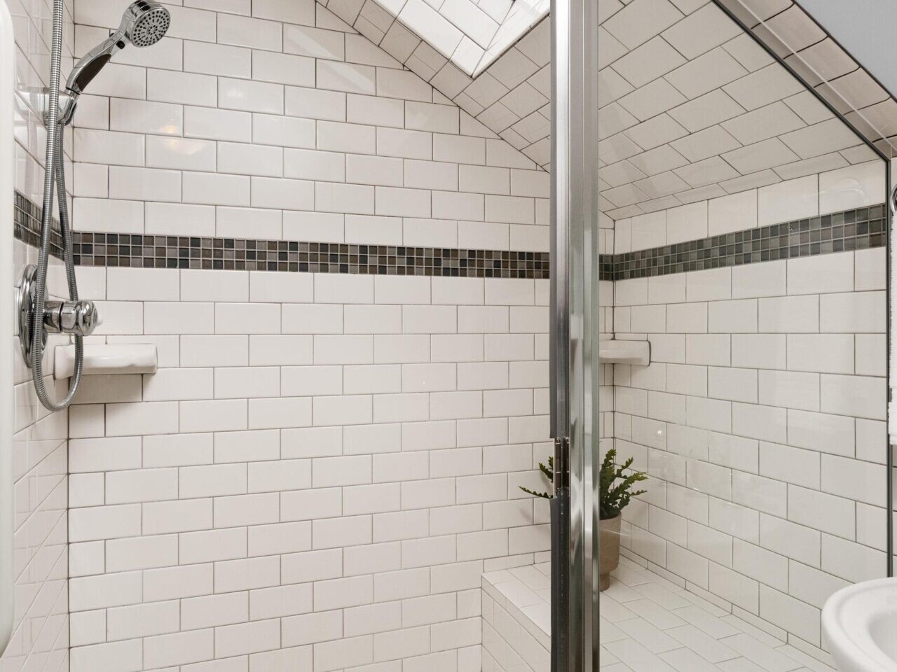 A modern bathroom features white subway tiles and a walk-in shower under a skylight. The shower has a glass door, a rainfall showerhead, and small hexagon tiles on the floor. There is a built-in bench with a small plant.