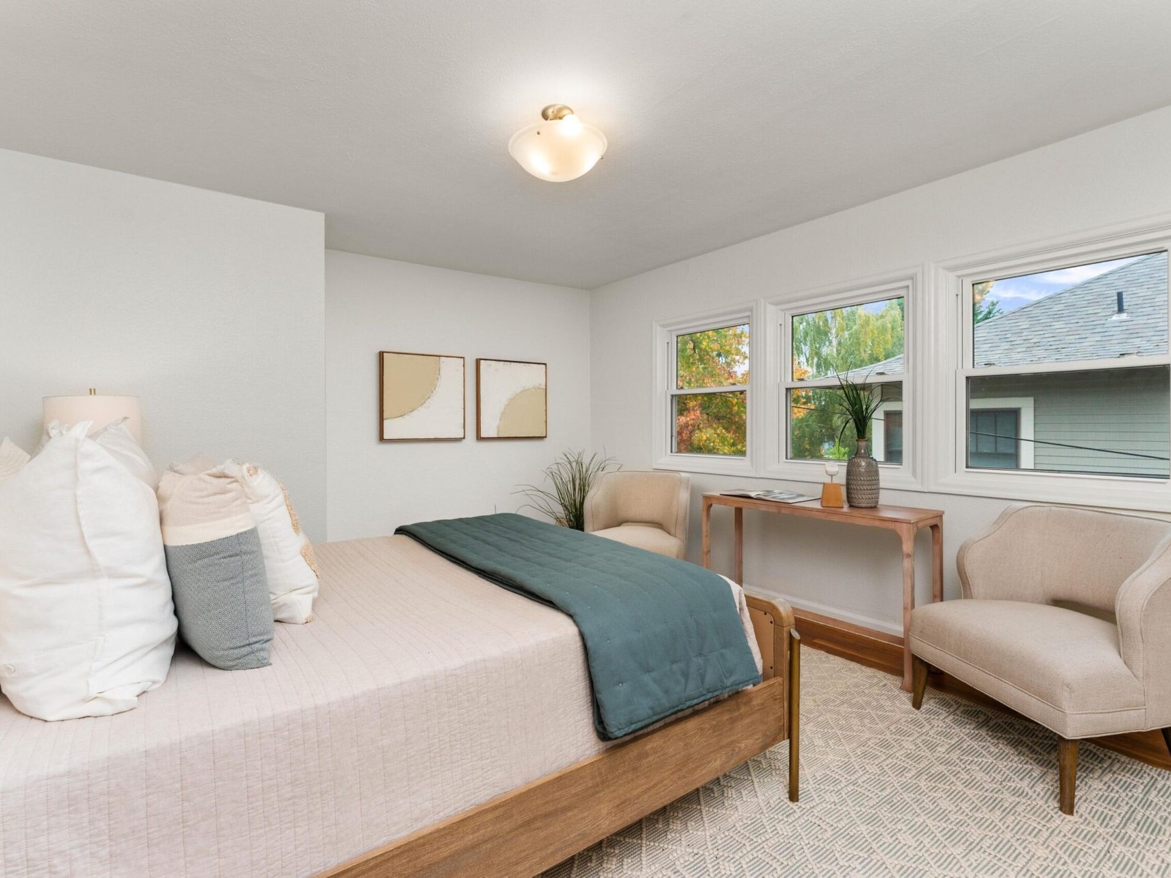 A cozy bedroom with a wooden bed, white and gray bedding, and a green blanket. Two beige abstract paintings hang on the wall. Theres a wooden desk with a chair by the window and a light beige armchair on a patterned rug.