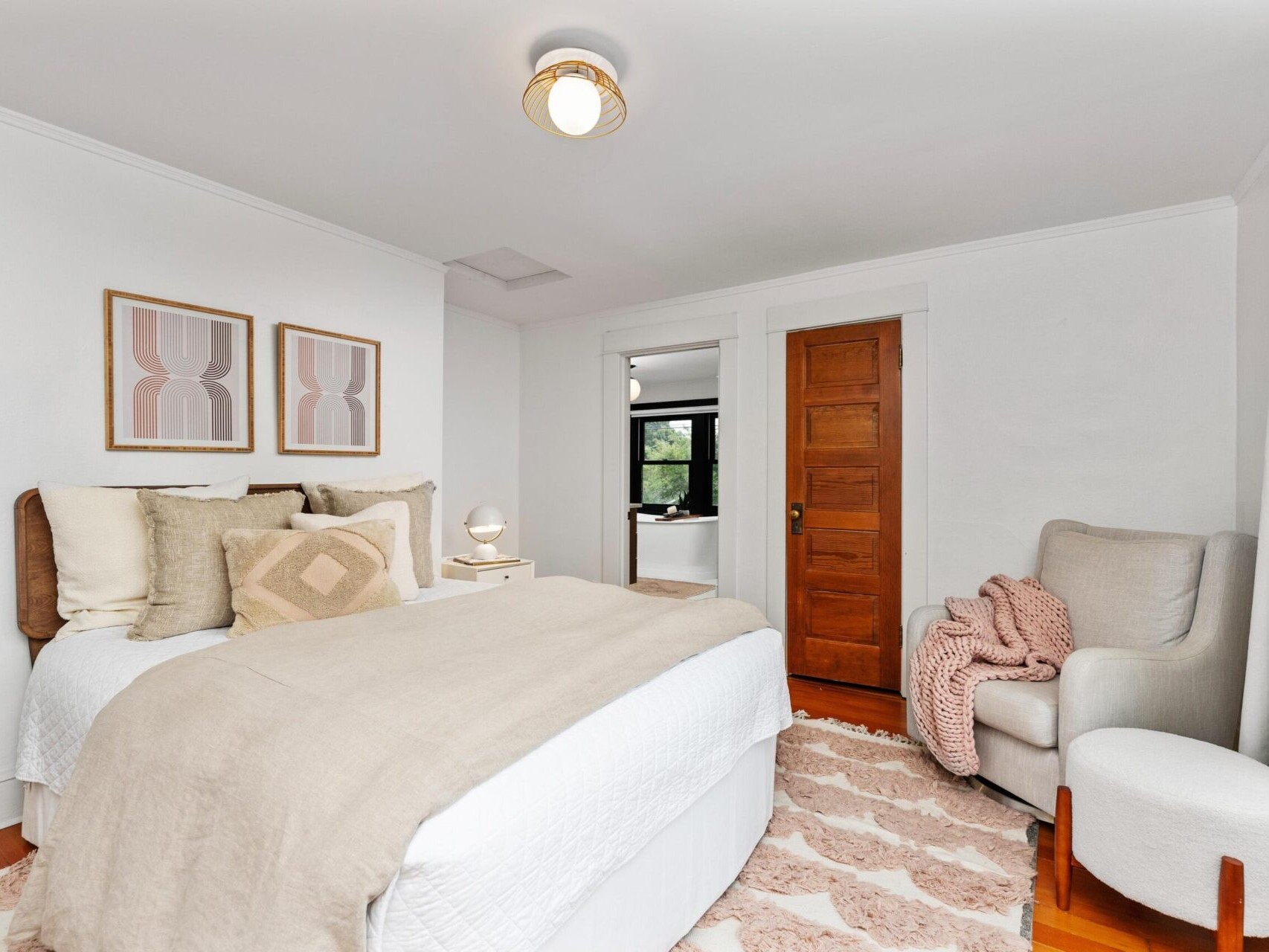 A cozy bedroom in Portland, Oregon, with a neatly made bed featuring beige and white bedding. Two abstract paintings hang above the bed. There's a plush armchair with a pink blanket and a small round stool. A wooden door and a large window are in the background.