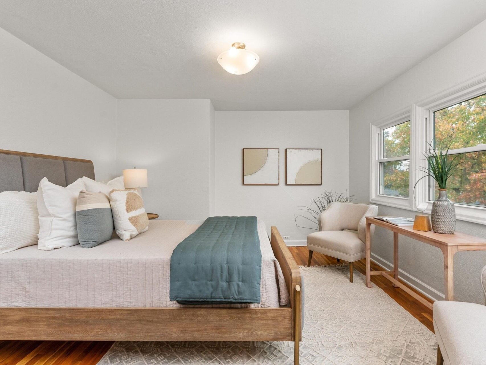 A cozy bedroom with a wooden bed, gray bedding, and multiple pillows. Two abstract paintings hang above the bed. The room features a light-colored rug, two beige chairs, a wooden desk, and a window with a view of greenery outside.
