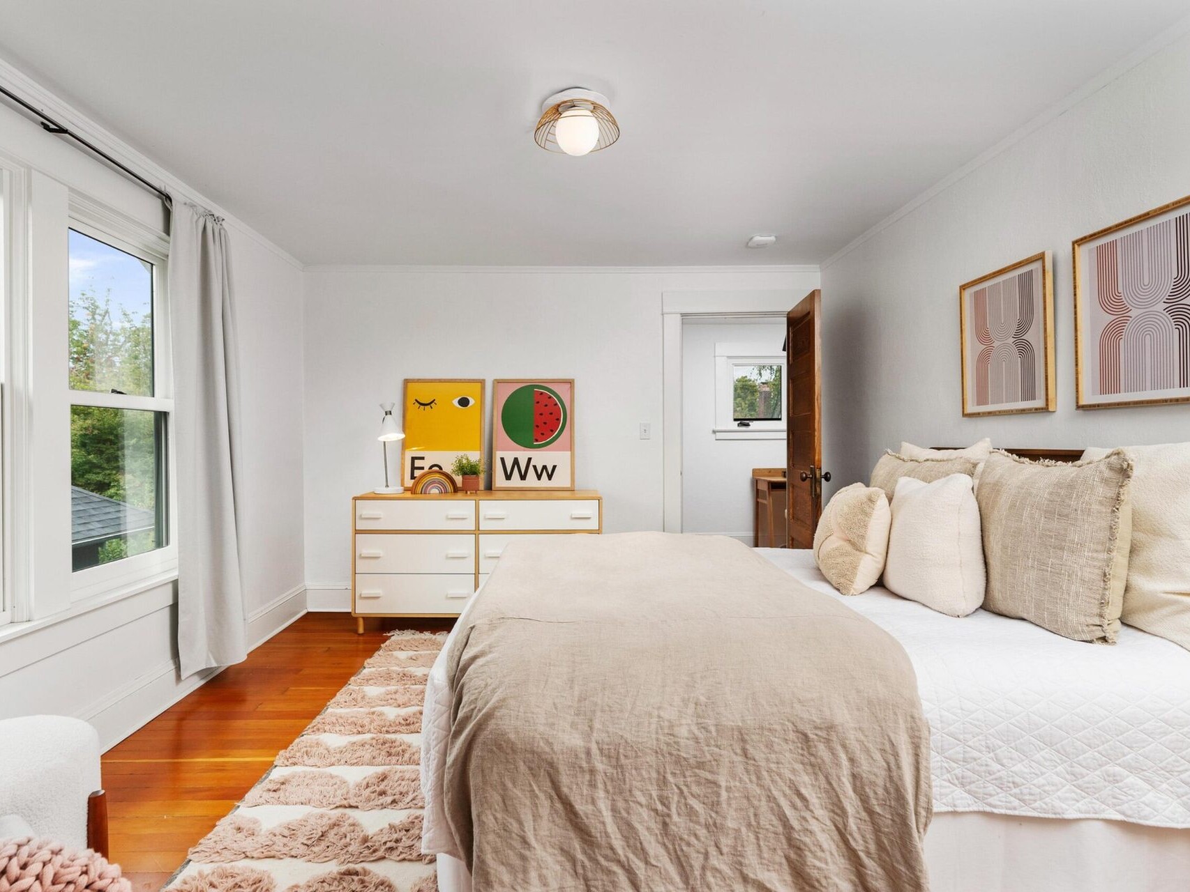 A cozy bedroom in a Portland, Oregon home, with a neatly made bed, beige bedding, and accent pillows. The room features two abstract wall art pieces, a dresser with colorful decor, a large window with white curtains, and a soft patterned rug on wooden floors.