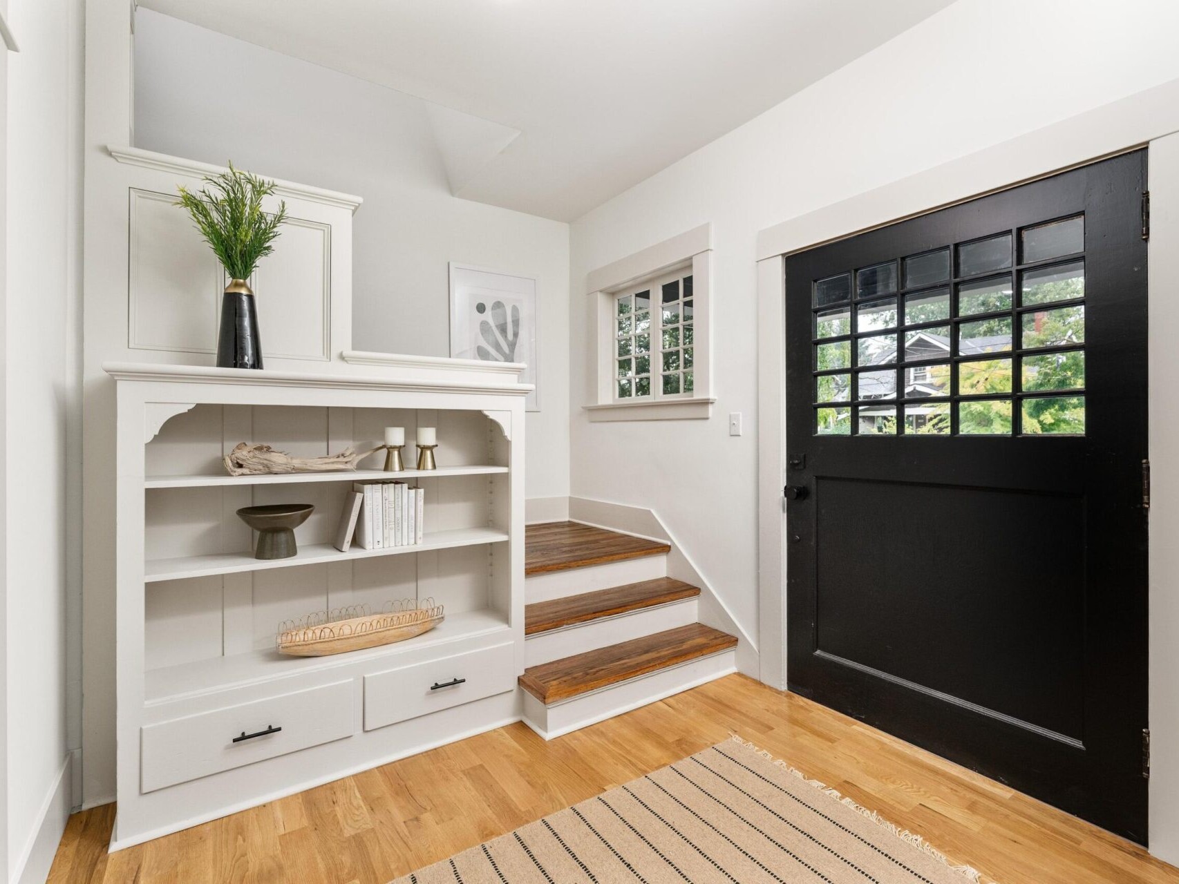 A cozy Portland, Oregon entryway features a black front door with grid windows, a white built-in bookshelf with decor, and wooden stairs leading up. The area boasts a light wood floor and a beige striped rug. A small window on the wall allows natural light in.