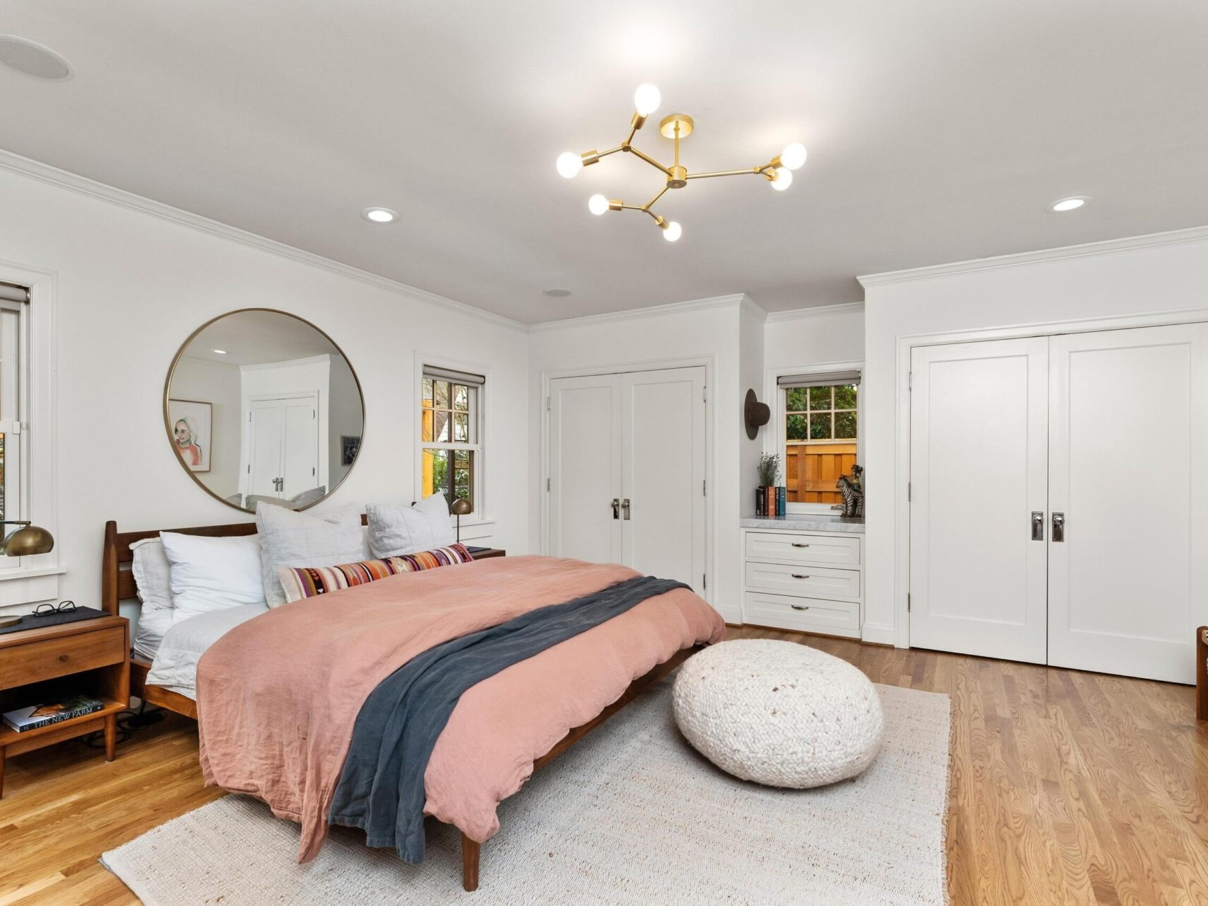 A modern bedroom in Portland real estate with a large bed featuring pink and gray bedding. A round mirror sits above the bed, beside a wooden nightstand. A pouf rests on a light rug, surrounded by white built-in closets, bright hardwood floors, and a geometric ceiling light.