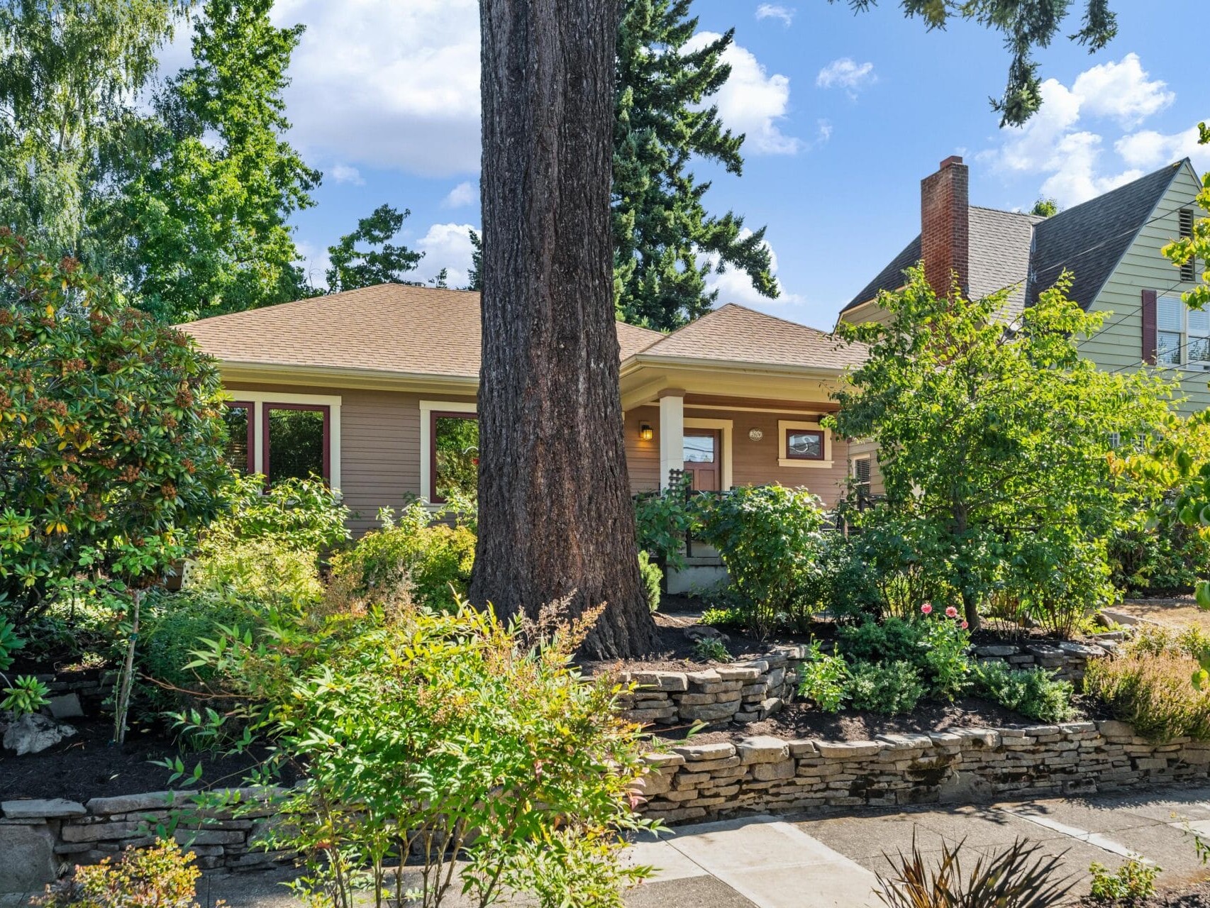 A quaint house with a tan exterior in Portland, Oregon, is partially obscured by lush greenery and a large tree. The yard is landscaped with various plants and shrubs, bordered by a stone wall. A bright, sunny day highlights the vibrant foliage.