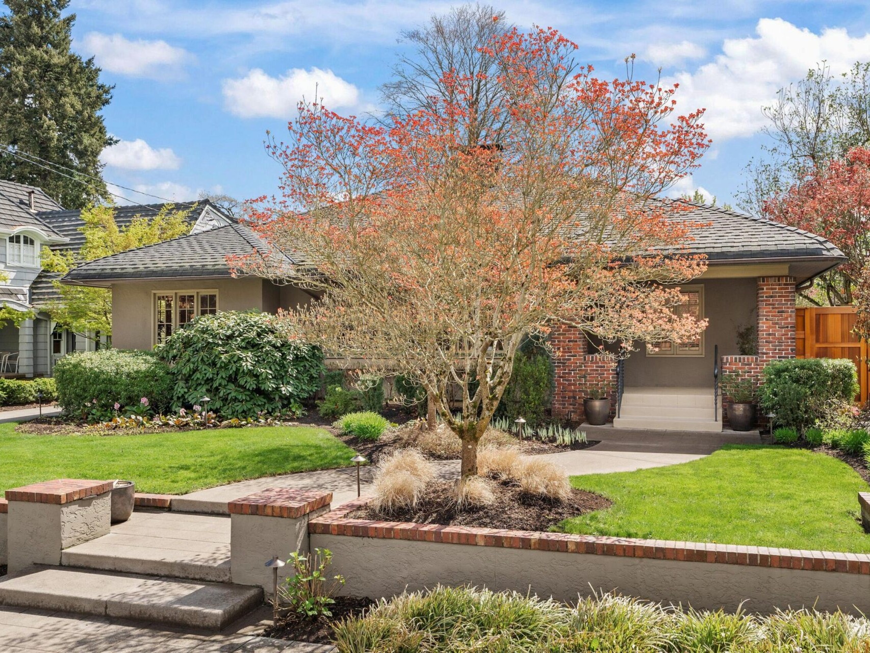 A suburban house in Portland Oregon real estate boasts a manicured front yard with a small tree adorned with red leaves, surrounded by lush green grass and bushes. The charming home features a brick facade, a sloping roof, and a concrete pathway welcoming you to the entrance.