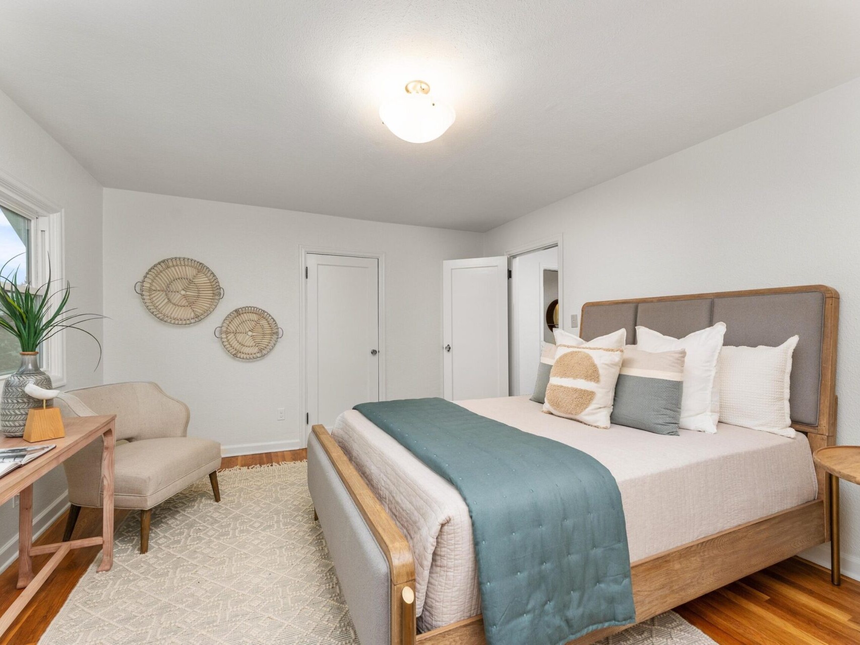 A modern bedroom with a wooden bed frame, beige and teal bedding, and a light gray area rug. Theres a beige armchair, a nightstand with a plant, and wall art. The room has two closed doors and a ceiling light.
