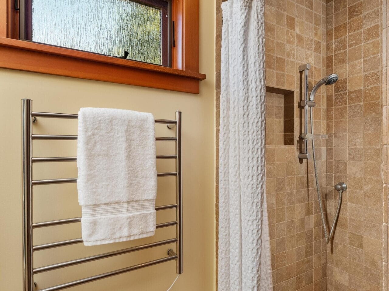 A bathroom in Portland, Oregon, features a beige tiled walk-in shower with a crisp white curtain. A towel hangs neatly on the heated rail to the left, while a high frosted window above adds an airy touch.
