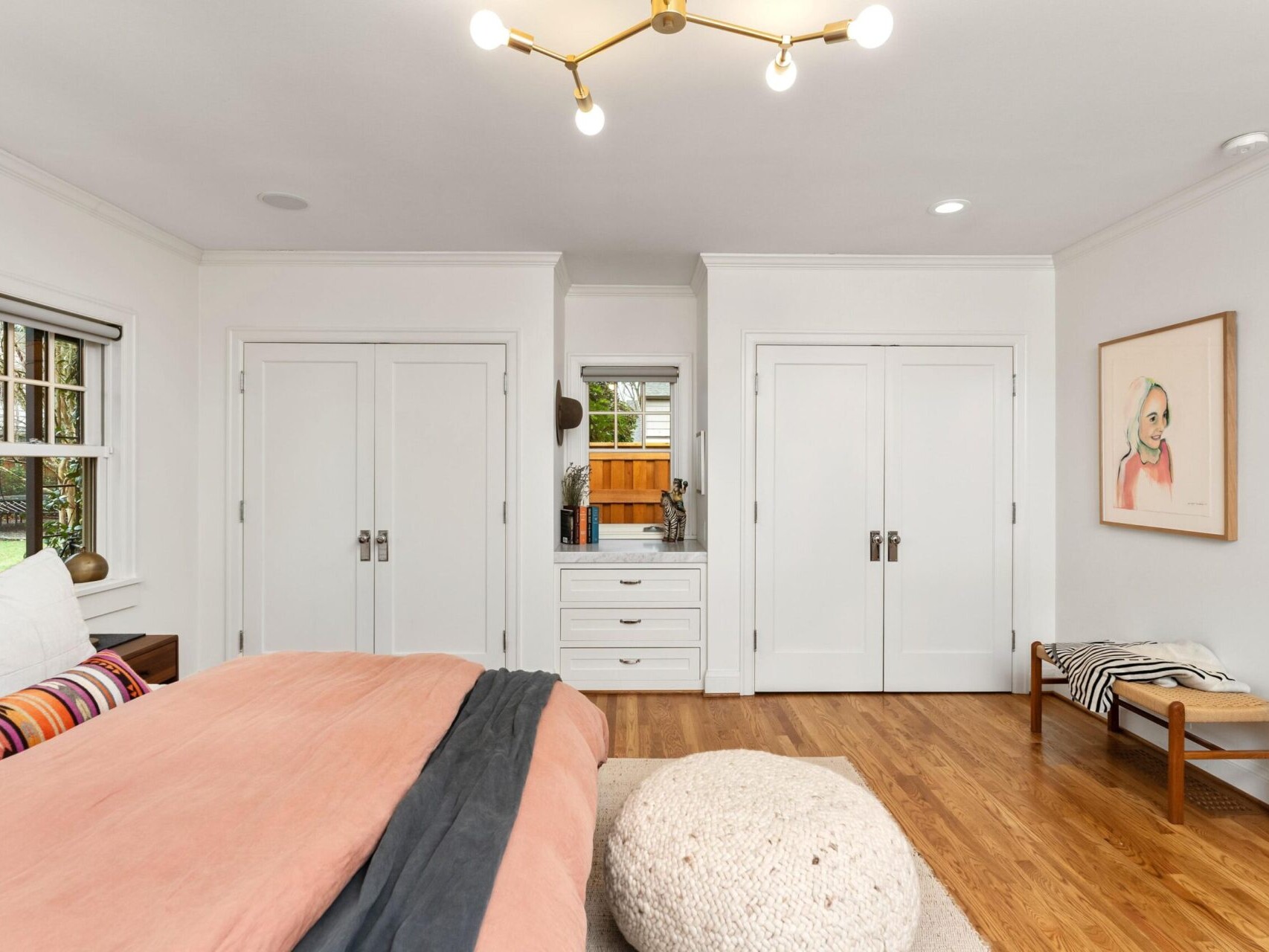 A cozy bedroom with a large bed covered in pink and gray bedding reflects Portland Oregon real estate charm. There's a soft pouf on the wooden floor. Two closets flank a central dresser with a mirror. A bench with striped fabric and framed artwork adorn the white wall, beneath a modern light fixture.