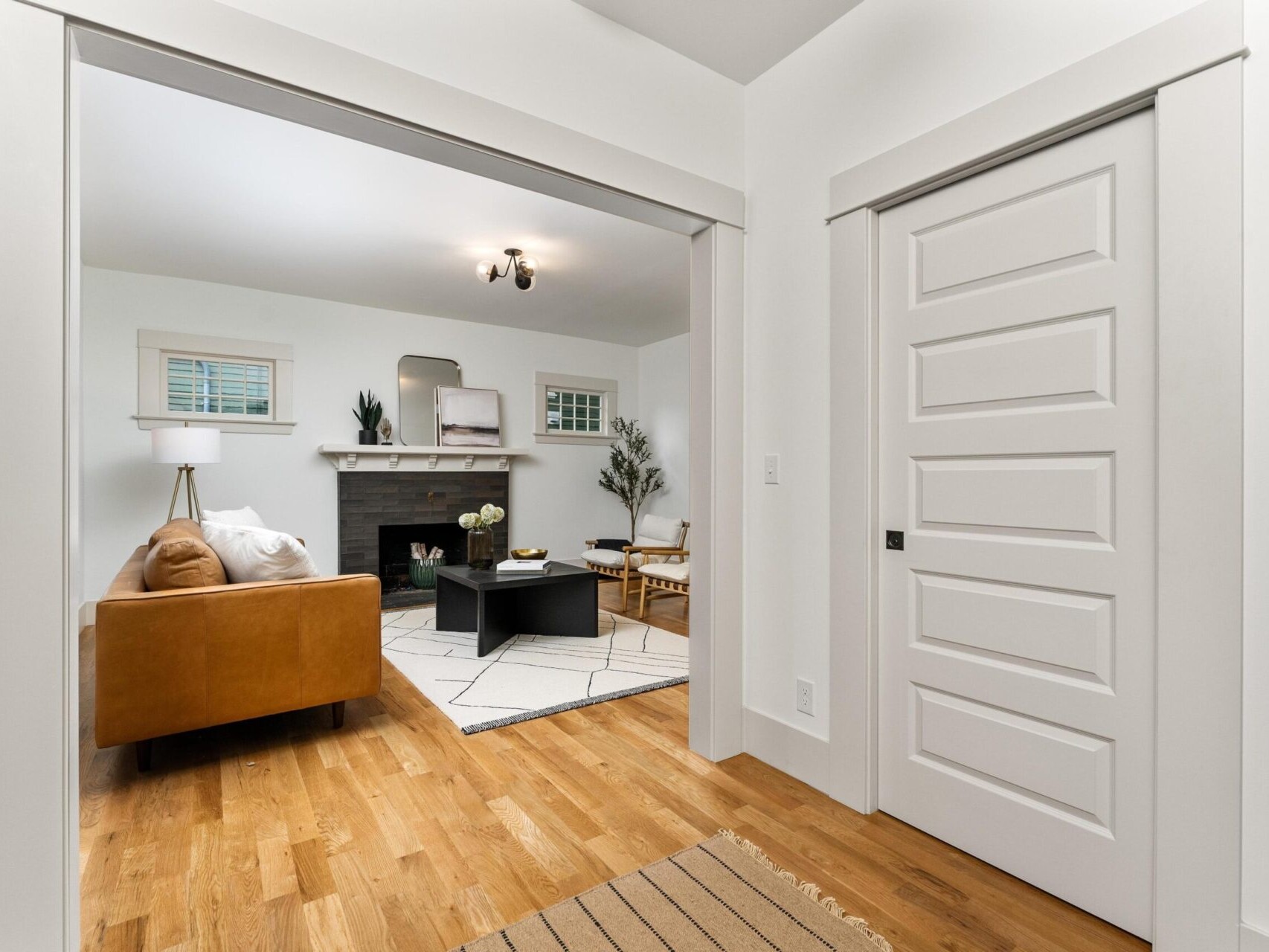 A cozy Portland, Oregon living room features white walls and hardwood floors. The space includes a brown leather sofa, geometric rug, fireplace with mirror above it, and potted plants. A doorway leads to this inviting area from a hallway adorned with a striped rug.
