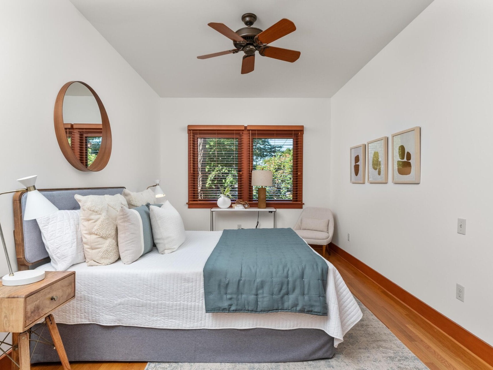 A minimalist Portland Oregon bedroom boasts a bed with white and teal bedding, a wooden side table, and a round mirror above the headboard. A chair rests in the corner beside a small desk with a lamp, two windows with wooden blinds, and art adorning the walls.