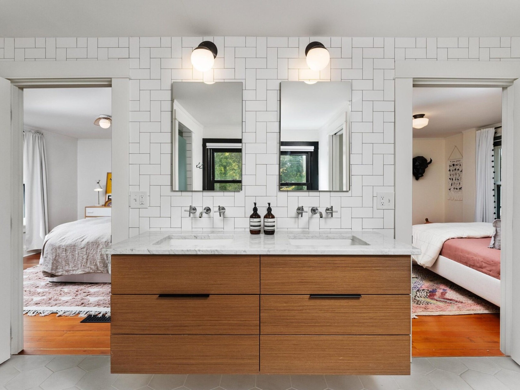 A modern Portland, Oregon bathroom features a double sink vanity with wooden drawers and two mirrors. Bright lighting fixtures hang above. Open doors lead to two separate bedrooms with beds and decor visible. The walls display a white tiled pattern.