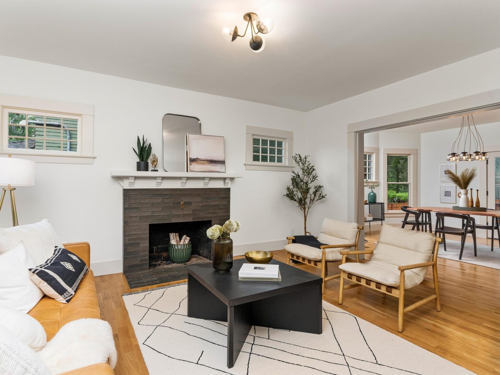 A modern living room in Portland, Oregon, features a brown leather sofa, two light wooden chairs, and a black coffee table on a geometric rug. A fireplace with decor and a large plant enhance the charm. In the background, there's a dining area with a wooden table and chairs.