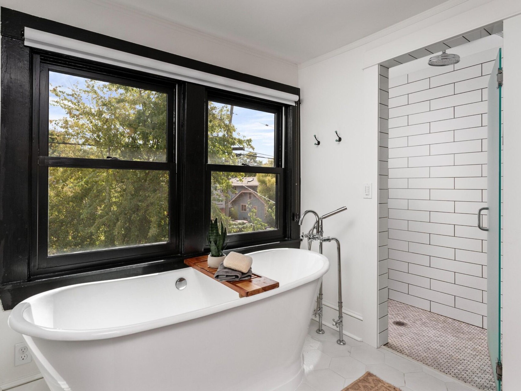 A modern bathroom in a Portland, Oregon home features a white freestanding bathtub with a wooden tray holding toiletries. The room has large windows offering tree views, a white subway tile shower to the right, and minimalist decor complemented by black window frames.