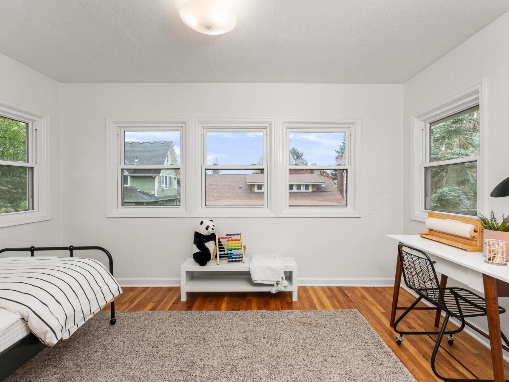 A bright bedroom with white walls, wooden flooring, and four windows. It features a bed with a striped blanket, a table with a lamp and plants, and a shelf with books and a panda toy. An area rug is centered on the floor.