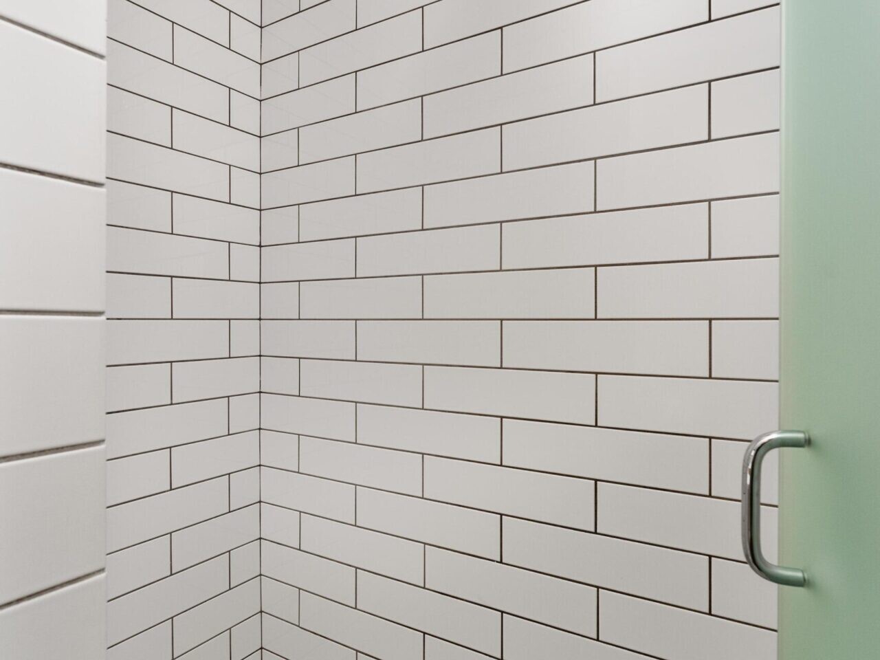 A minimalist shower in Portland, Oregon features sleek white rectangular subway tiles on the walls and small mosaic tiles on the floor. A glass door with a metal handle is partially visible on the right side, adding a touch of elegance to its serene design.