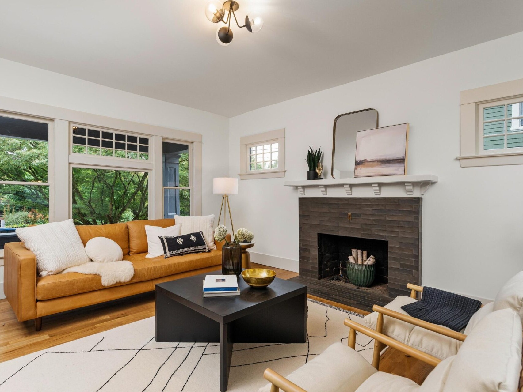 A cozy living room in Portland, Oregon, showcases a brown leather sofa and two beige armchairs around a black coffee table. It features a black brick fireplace, a geometric rug, and a large window with views of the lush greenery outside. A modern light fixture hangs from the ceiling.