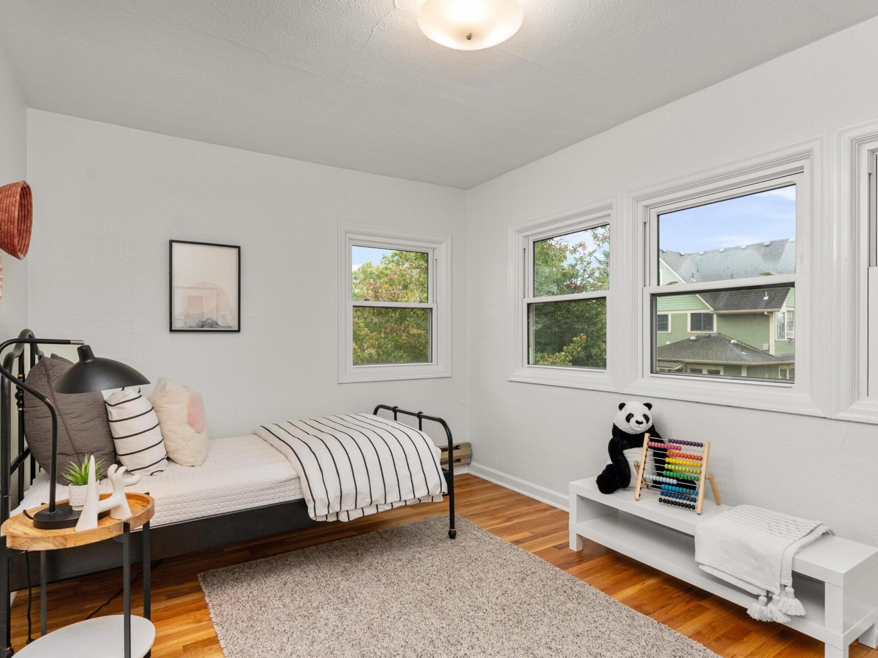 A cozy bedroom with a single bed and striped bedding. A small table holds a lamp and plant. A shelf with a plush panda, abacus, and a blanket is by the window. Three windows let in natural light, revealing a leafy neighborhood. A rug covers the wooden floor.