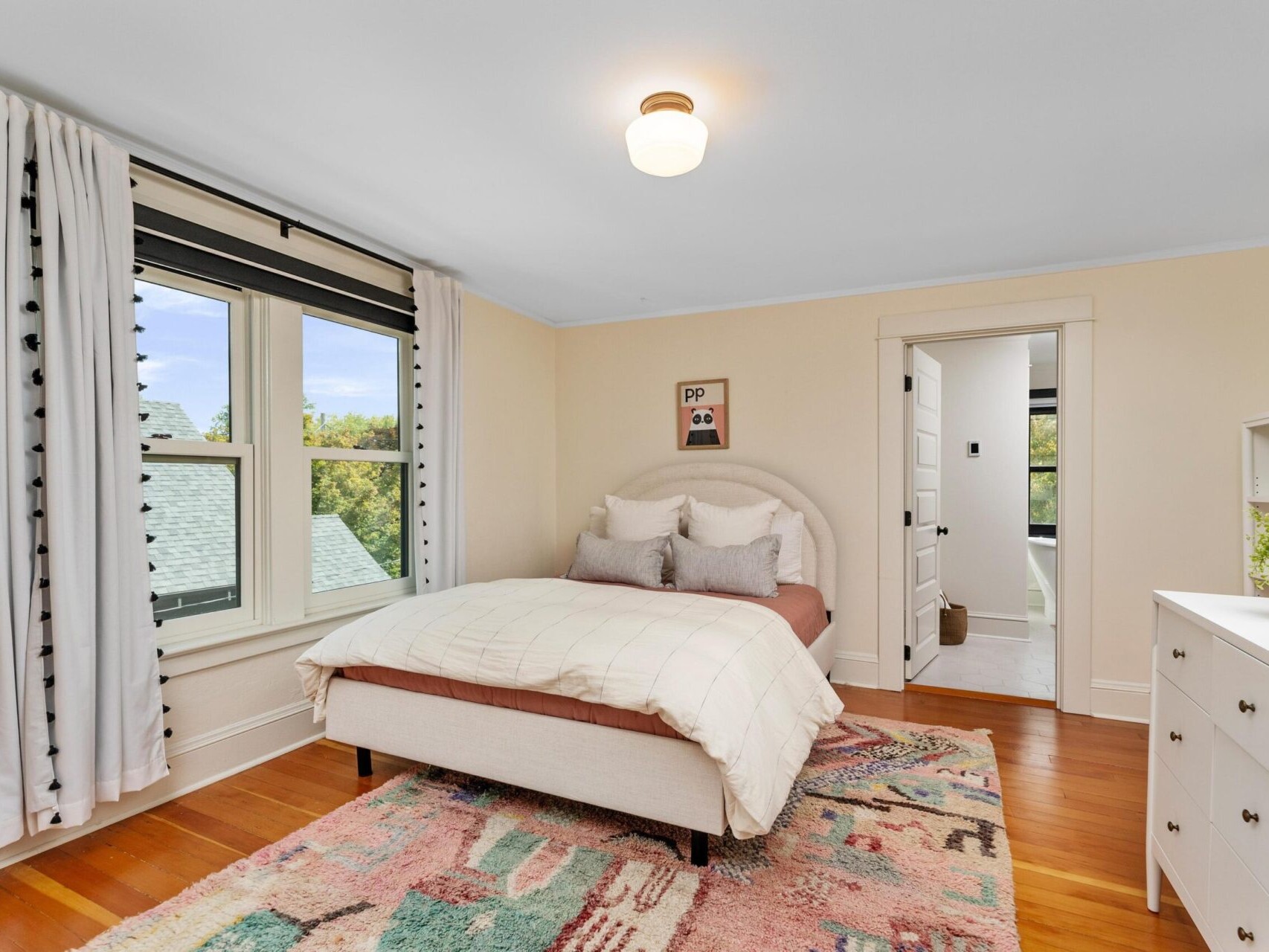 A cozy Portland, Oregon bedroom features a bed with a beige headboard and neutral bedding, complemented by a colorful rug on wooden floors. A white dresser adorned with plants sits near large windows with light curtains, while a doorway opens to reveal another room with an outdoor view.