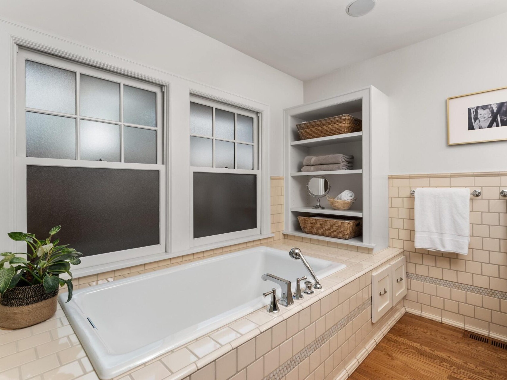 A beautiful bathroom design ideal for a Portland Oregon real estate listing, featuring a white rectangular bathtub with silver fixtures against cream-tiled walls. A shelf with towels and baskets is next to the tub, beneath two frosted windows, with a sleek black-and-white photo complementing the space.