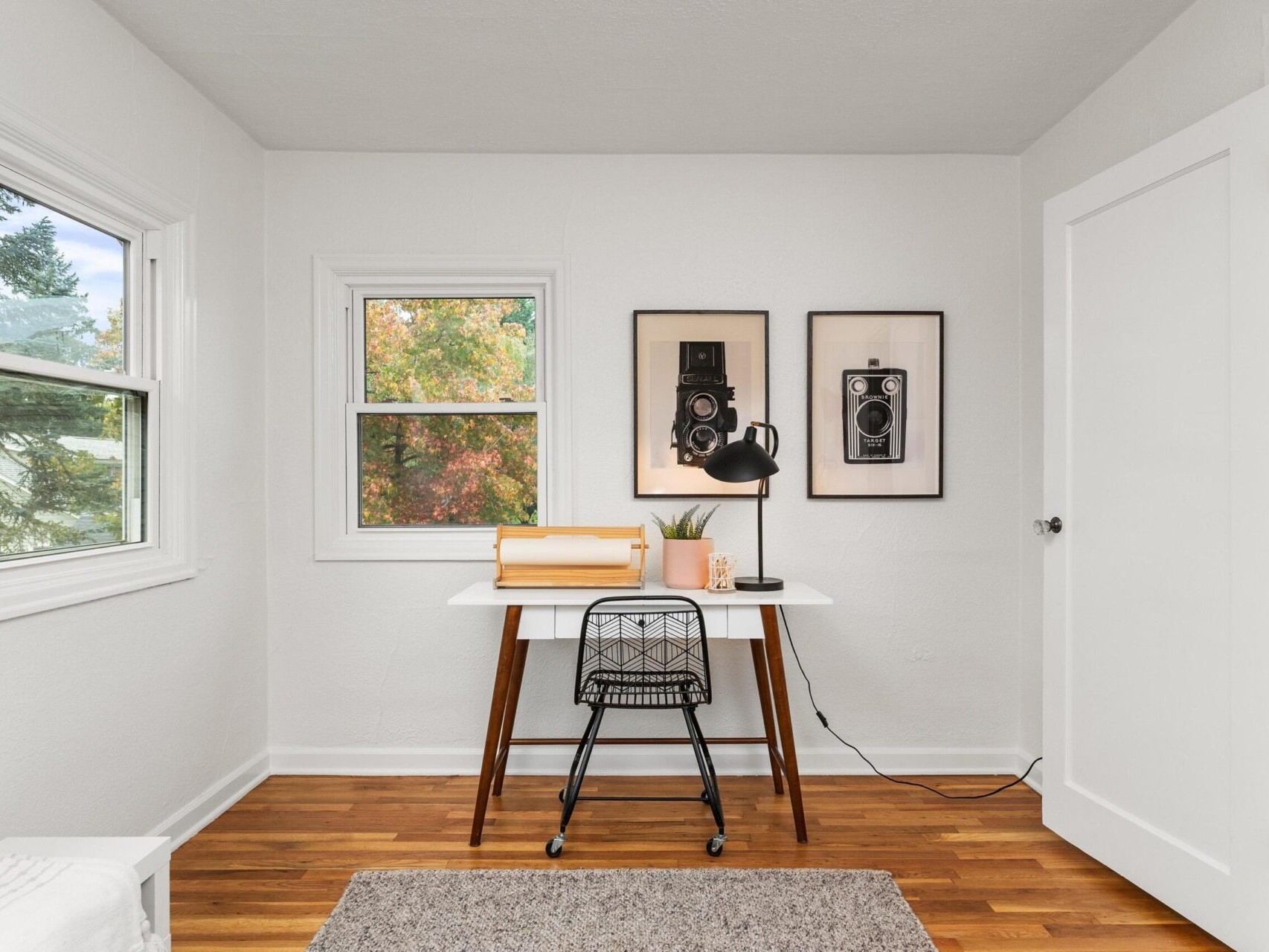 Minimalist home office with a small wooden desk and black chair facing two windows. A black lamp, potted plant, and decorative boxes are on the desk. Two framed camera illustrations hang above, and a lush view of trees is visible outside.