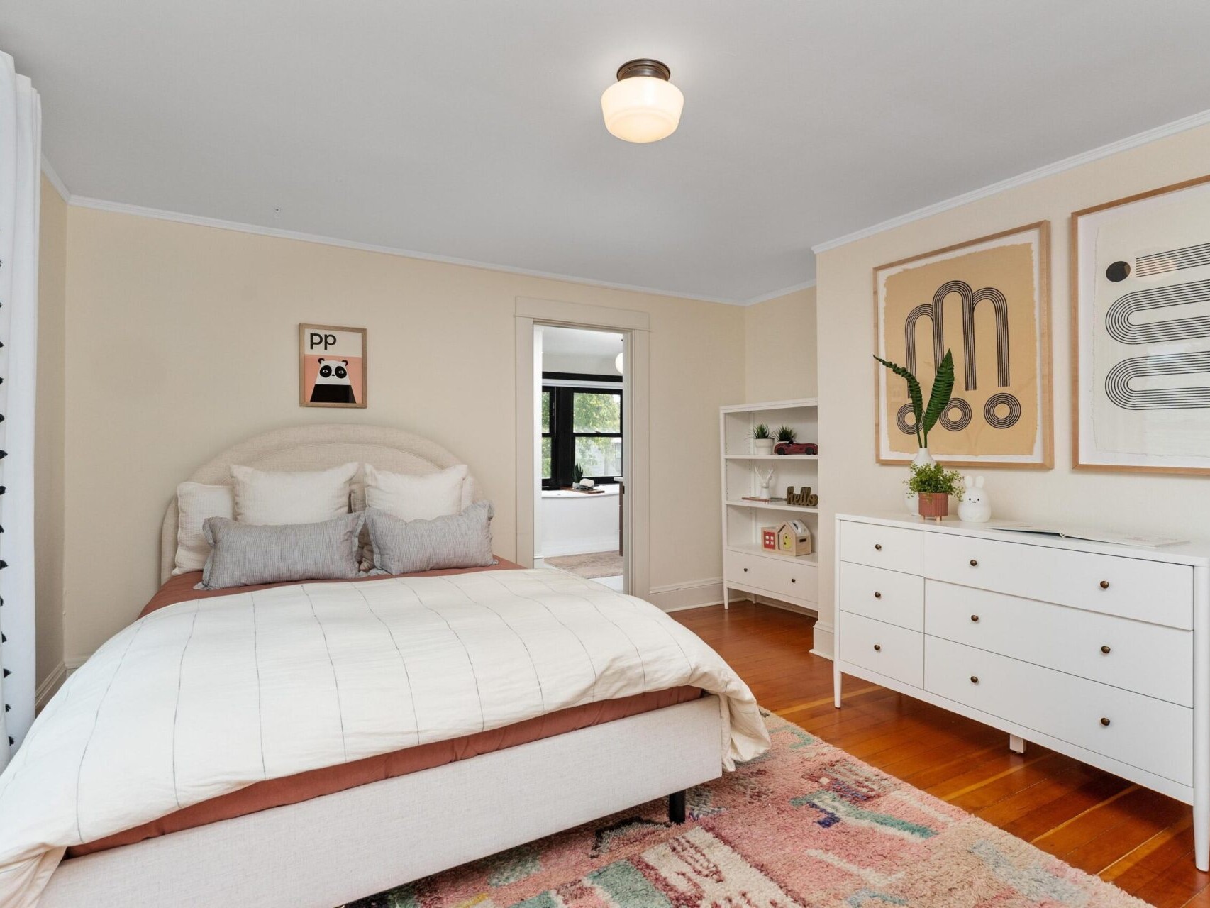 A cozy bedroom in Portland, Oregon, featuring a neatly made bed with neutral-toned bedding and colorful pillows. A white dresser sits beneath framed abstract art, alongside a bookshelf. Patterned curtains adorn the window, while warm wooden floors and a soft rug complete the inviting space.