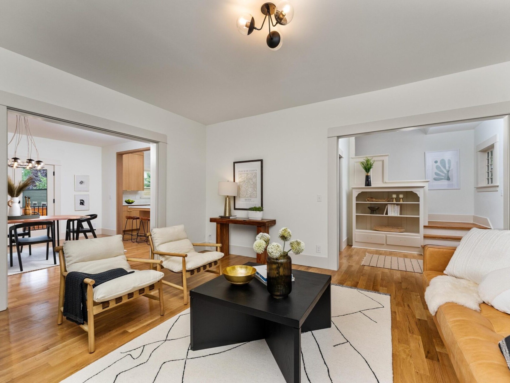 A modern living room in Portland, Oregon features a leather sofa, two wooden chairs, and a black coffee table on a patterned rug. The room opens to a dining area and kitchen. Neutral tones and minimalist decor create a cozy atmosphere, with a staircase visible in the background.