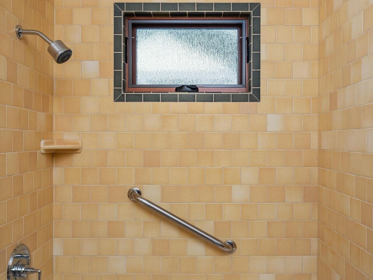 A bathroom in Portland, Oregon, showcases a white bathtub with a silver faucet and wall-mounted showerhead. The yellow tiled walls include a textured window with a dark frame and a safety grab bar installed horizontally. A soap dish is conveniently attached to the wall.