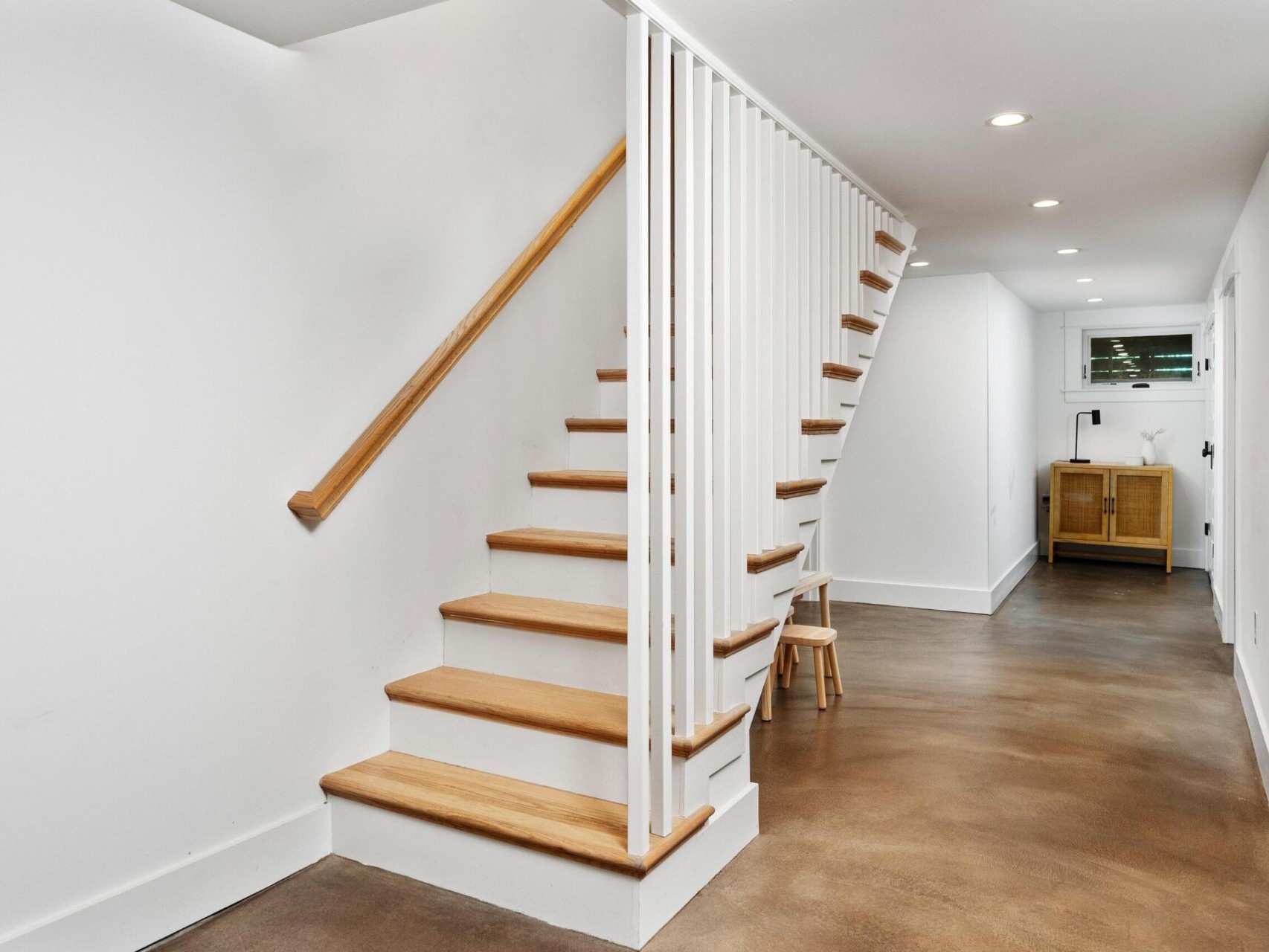 A modern, minimalist hallway in Portland, Oregon, features a wooden staircase with white railings. The space boasts polished concrete flooring and pristine white walls. A small wooden stool and a cabinet with an open shelf add functional charm to the serene backdrop.