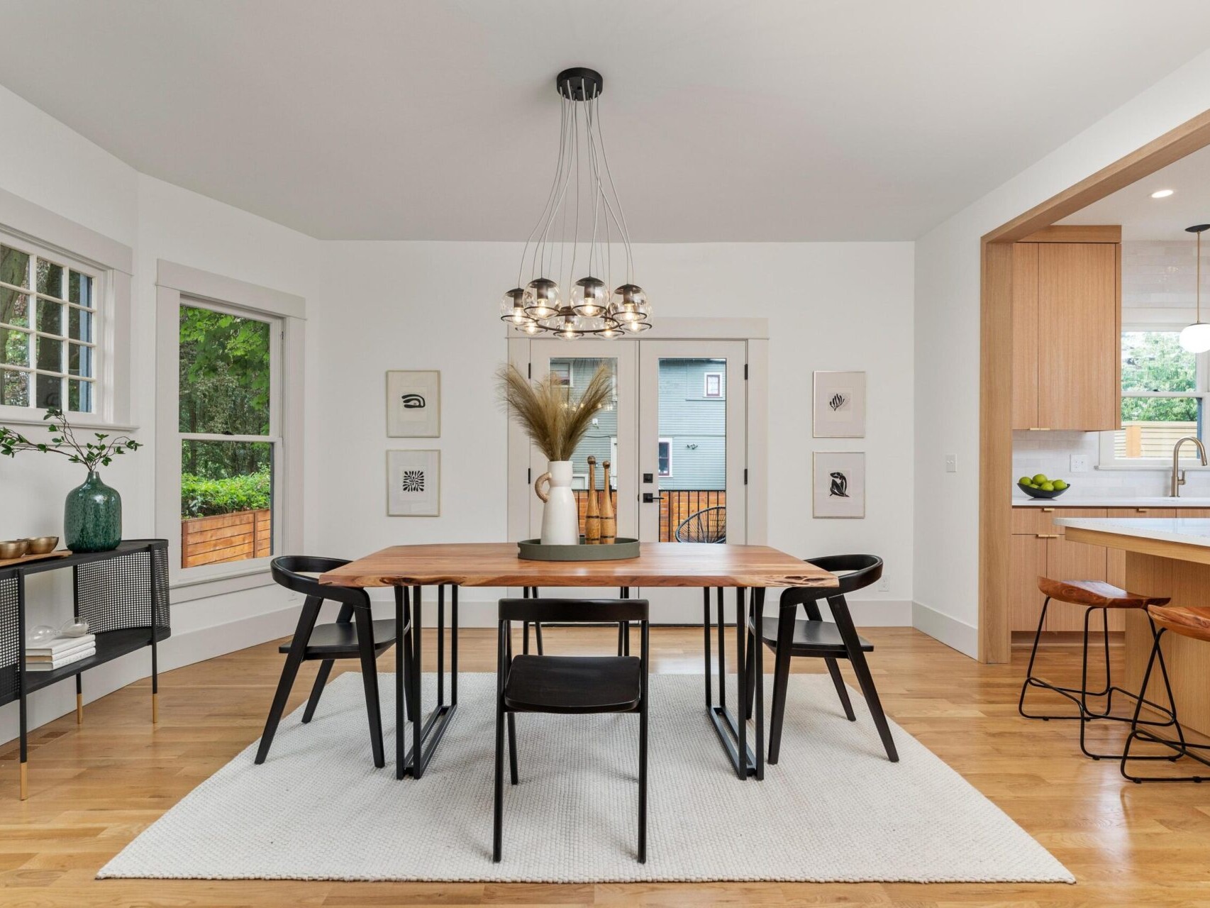 A modern dining room in Portland, Oregon, features a wooden table, six black chairs, and a central chandelier. The room boasts white walls, windows with scenic views, wall art, and an adjacent kitchen with bar stools and light wood cabinetry. A beige rug graces the wooden floor.