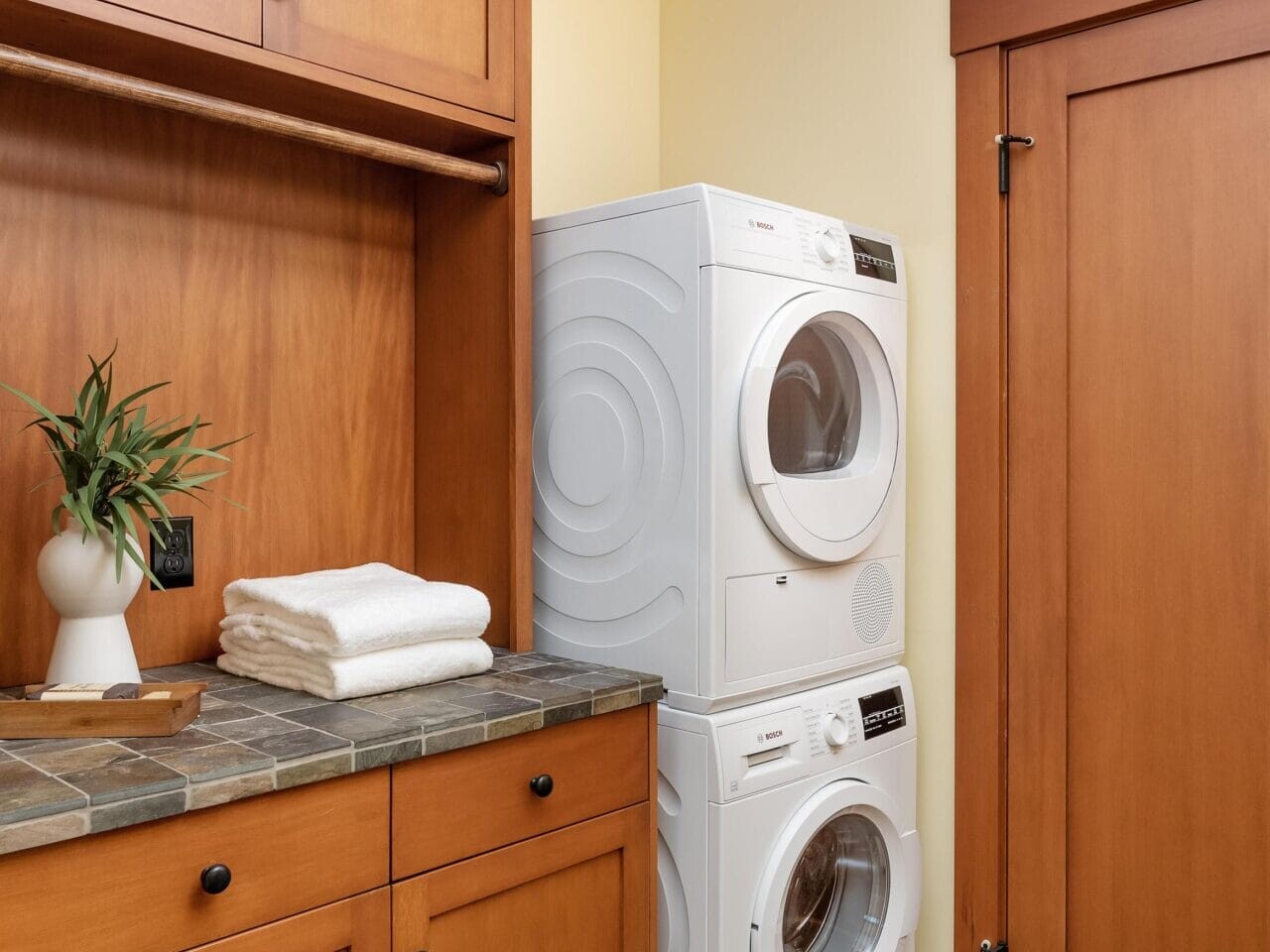 A cozy Portland, Oregon laundry room with a stacked washer and dryer beside wooden cabinets. A small potted plant and folded white towels adorn the countertop. The space is completed with a tiled floor and a rustic wooden door.