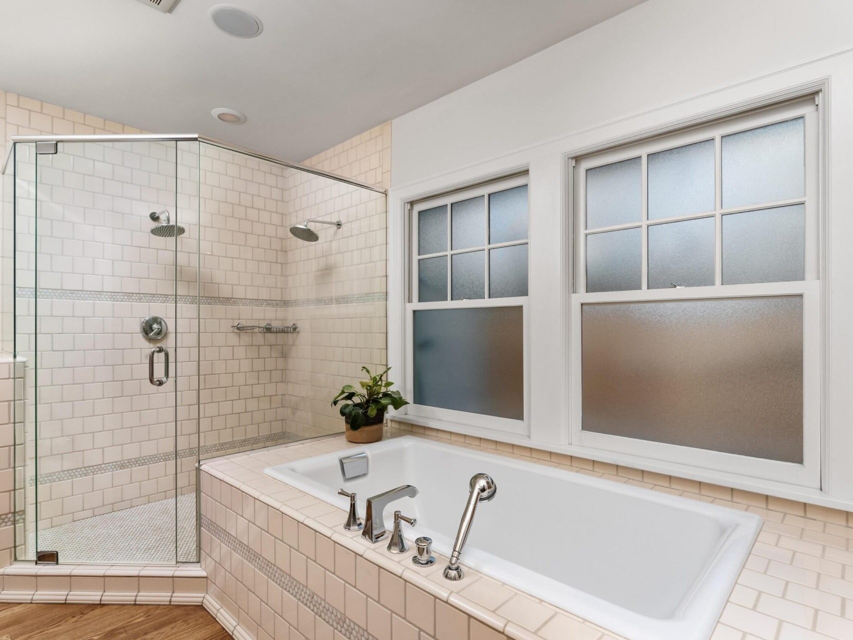A bathroom in a Portland Oregon Real Estate listing features a glass-enclosed corner shower with dual showerheads and a built-in shelf. Adjacent is a white tiled bathtub with chrome fixtures. Two frosted windows provide natural light, while a small plant decorates the tub corner.