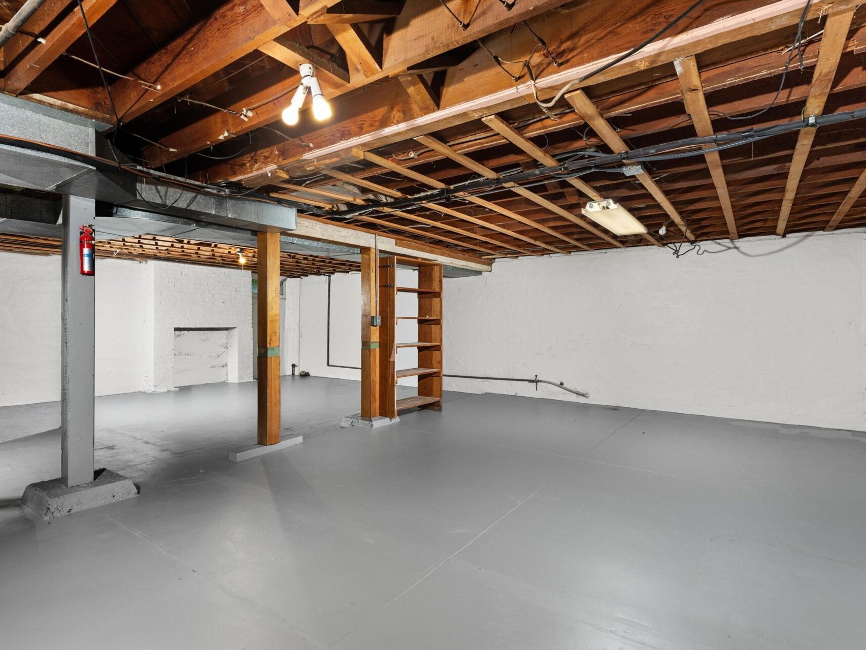 A spacious basement with unfinished wooden beams and exposed pipes. The floor is painted gray, and the walls are white. A ladder-like wooden shelf leans against one wall, and light bulbs hang from the ceiling, illuminating the area.