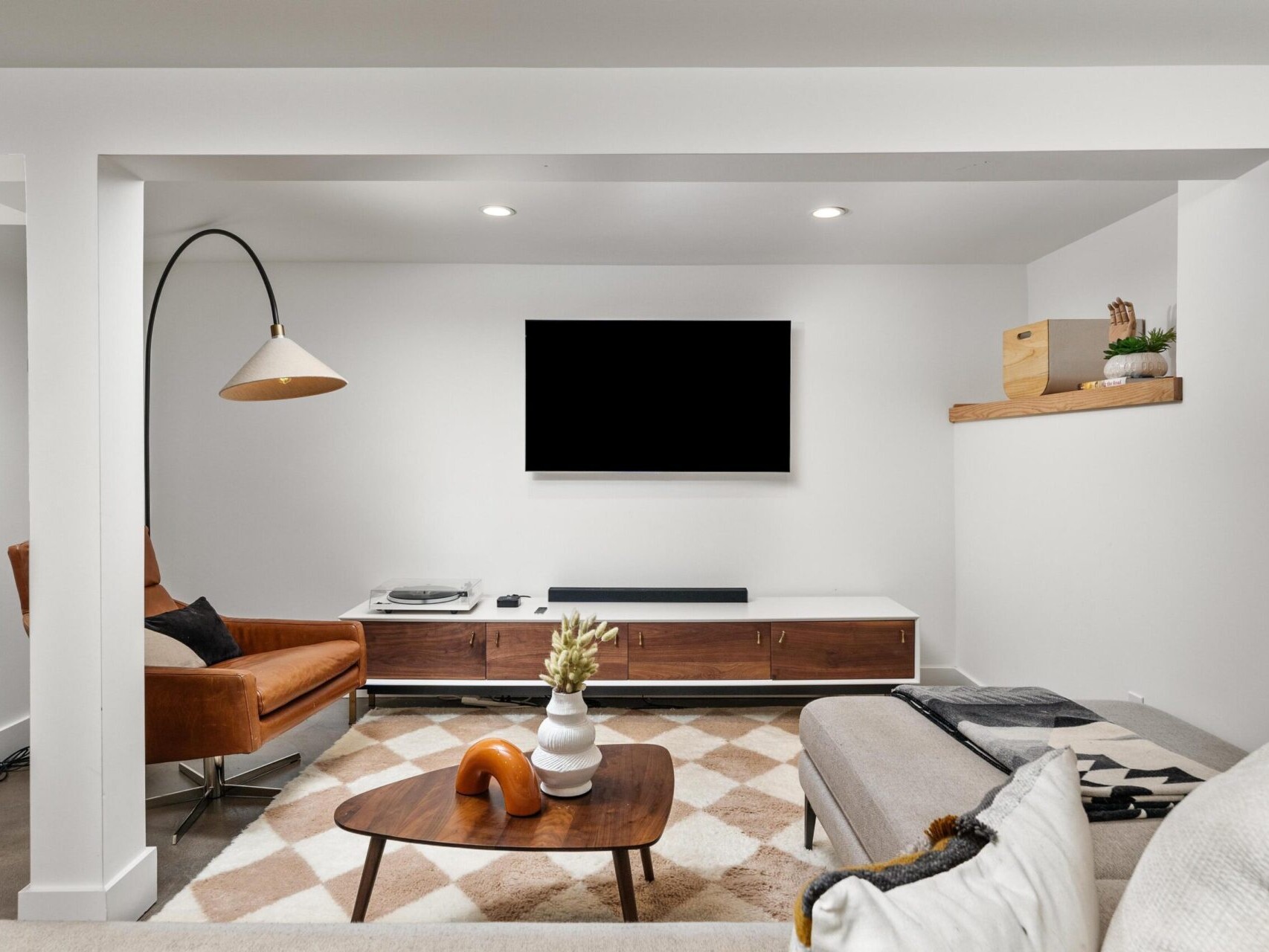 A modern living room in Portland, Oregon features a wall-mounted TV, a brown leather armchair with a floor lamp, and a beige sofa. A geometric-patterned rug covers the floor with a wooden coffee table at the center. Shelves hold decorative items.