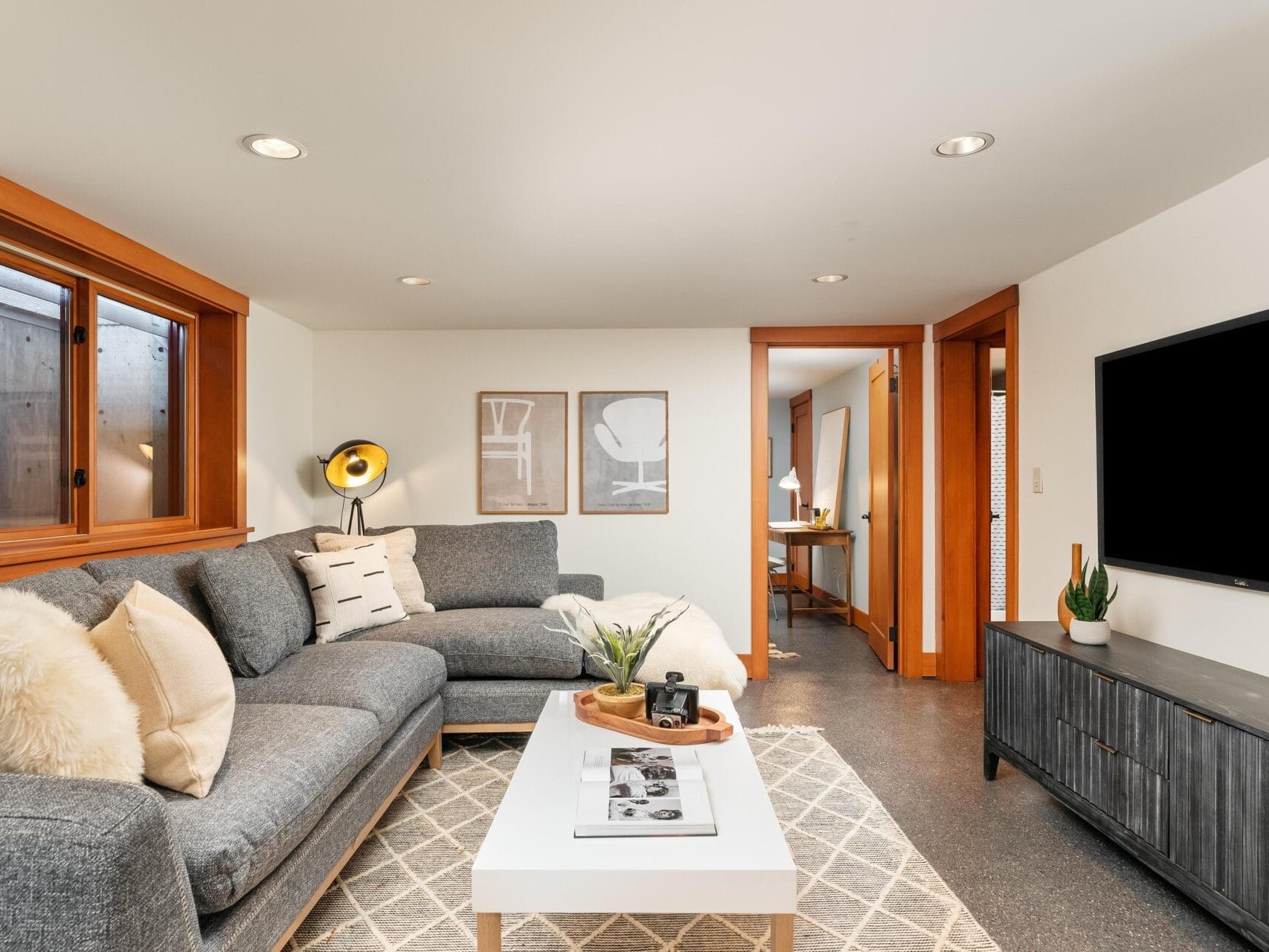 A cozy Portland, Oregon living room features a gray sectional sofa adorned with white and patterned cushions. A white coffee table displays magazines and a plant, while a large flat-screen TV is mounted on the wall. Warm wooden accents frame the windows and doorways, enhancing the inviting atmosphere.