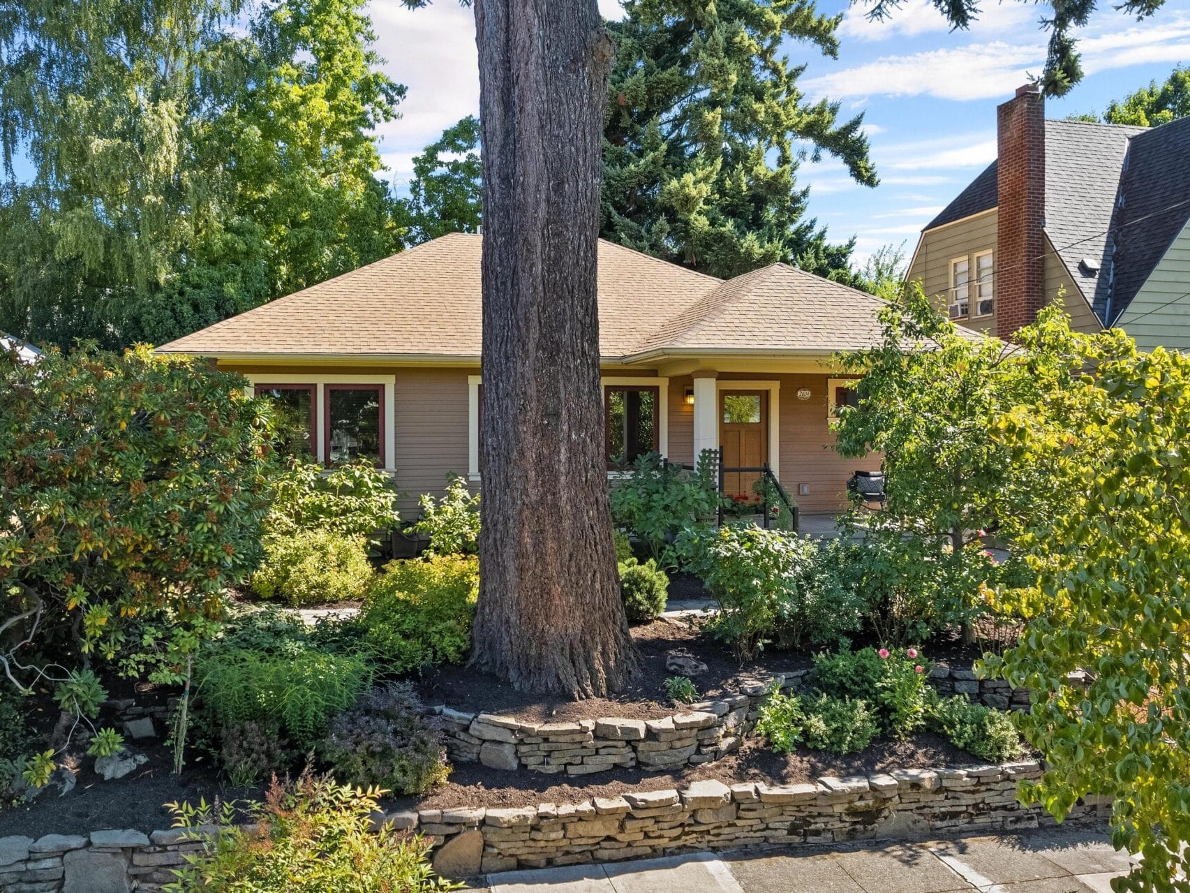A charming house with a beige facade is nestled among Portland, Oregon's lush greenery and trees. A large tree stands prominently in the front yard, surrounded by well-maintained landscaping with flower beds and a stone wall. The sky above this Portland gem is blue with scattered clouds.