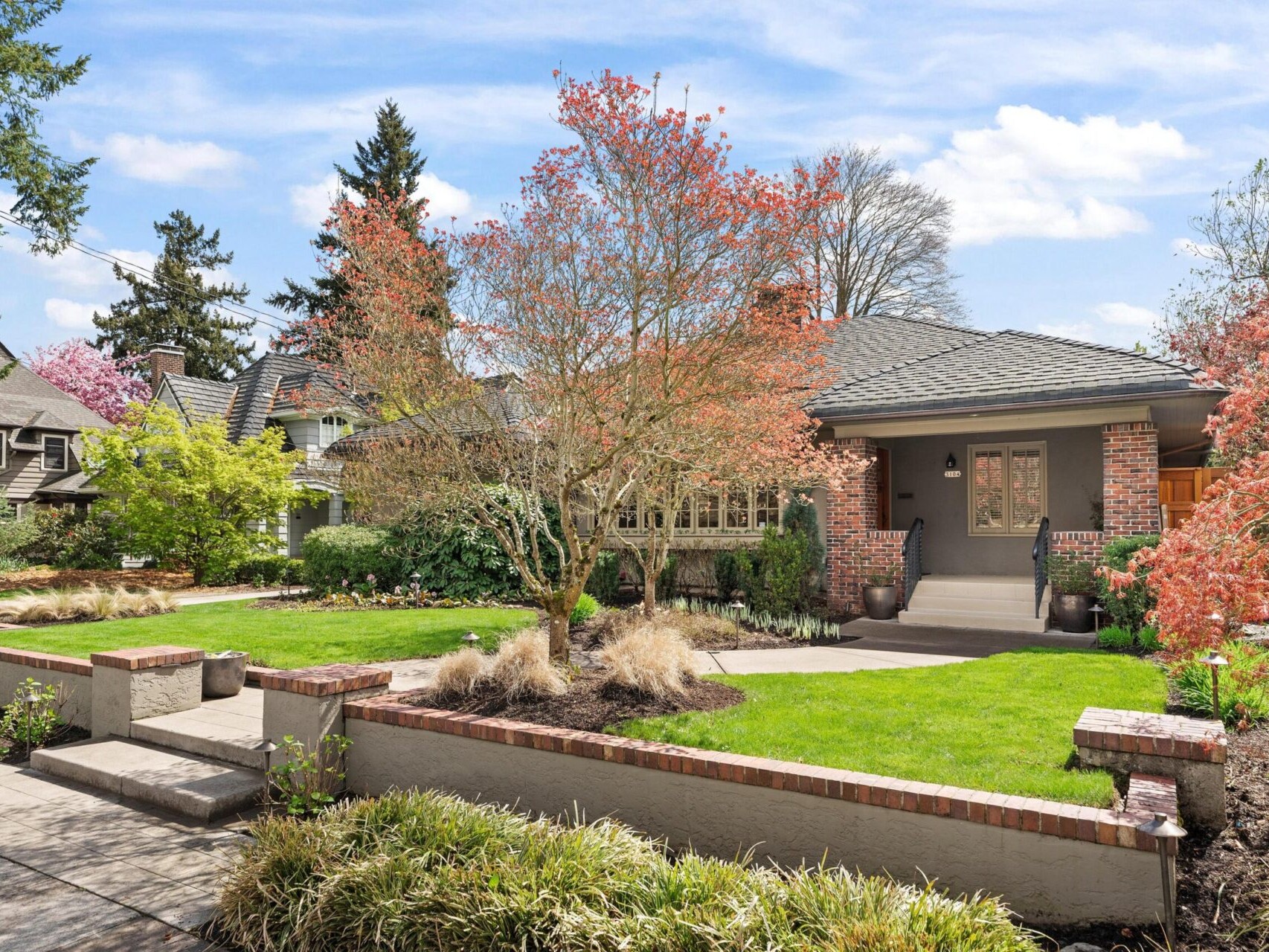 Charming brick house with a gabled roof and covered porch, nestled among lush landscaping in a quiet Portland neighborhood. A tree with reddish leaves graces the well-maintained front yard, and the sky is blue with fluffy clouds—a gem in Portland Oregon real estate.