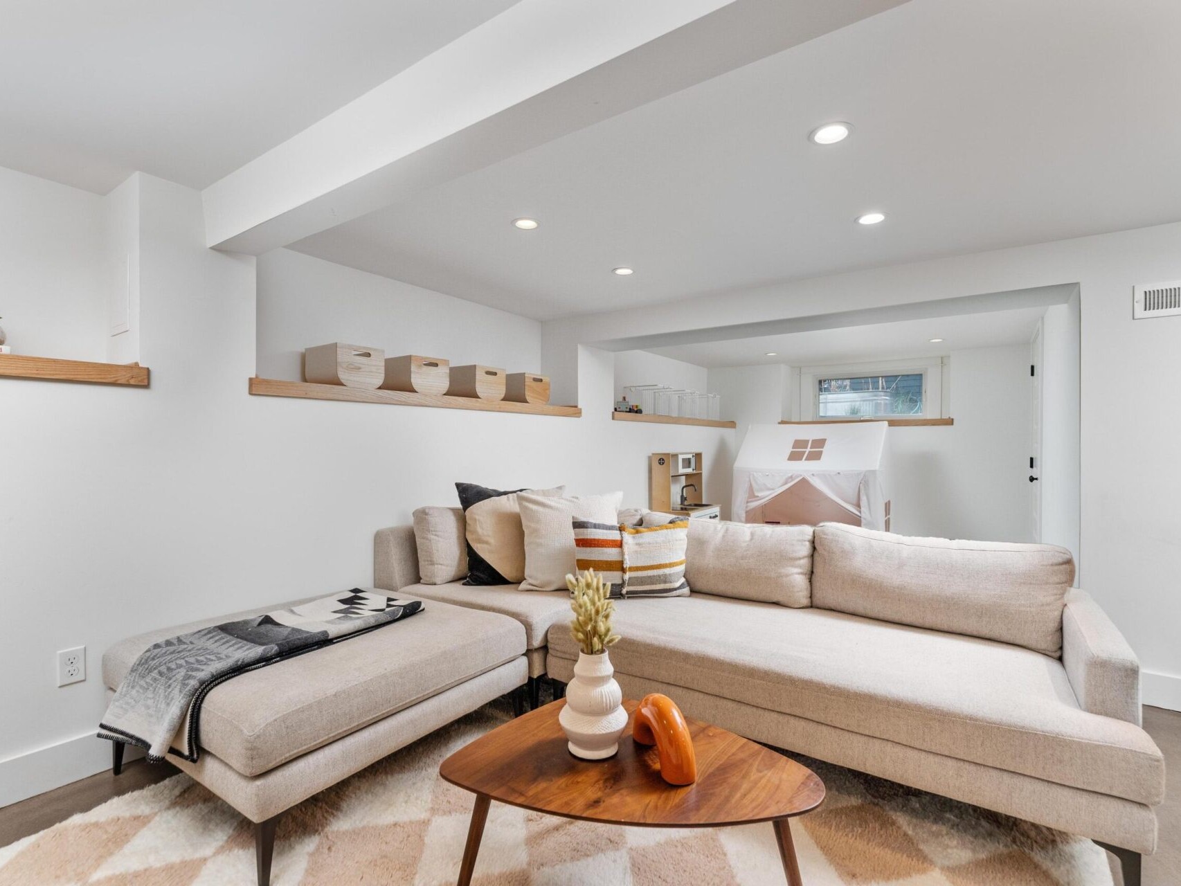 A cozy Portland, Oregon living room features a light beige sectional sofa, a wooden coffee table, and a geometric-patterned rug. Shelves with baskets and small decor items adorn the walls, adding warmth to the bright and spacious space.