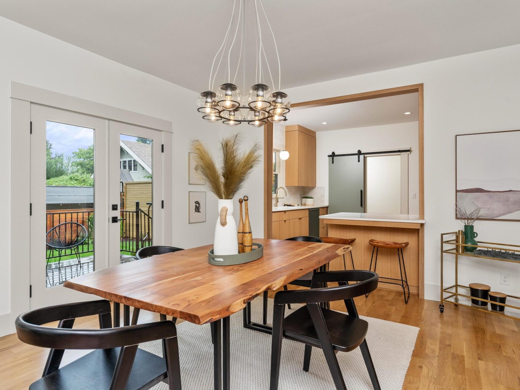 Modern dining room with a Portland, Oregon vibe features a wooden table, black chairs, and pendant light. Large glass doors open to a garden. Minimalist decor includes pampas grass in a vase and framed art. The kitchen is visible in the background through a wide doorway.