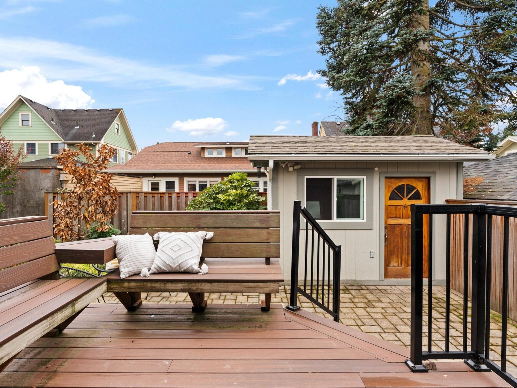 A backyard scene with wooden decking and benches, featuring two decorative cushions. A small shed with a wooden door and window is in the background, surrounded by a brick patio. There are houses and a tree visible beyond the fence.