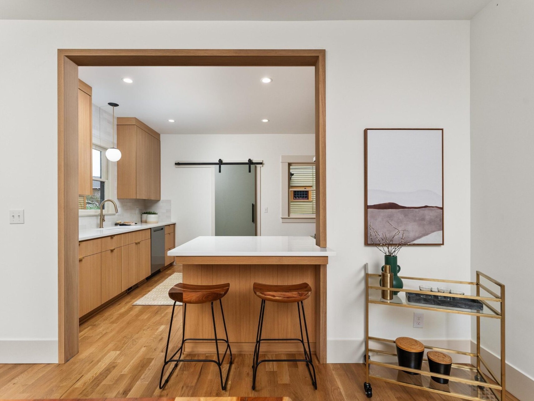 A modern Portland, Oregon kitchen features light wood cabinetry and a white countertop island. Two wooden bar stools are placed at the island. The room includes a decorative cart, framed artwork, and a sliding door leading to an adjacent area.