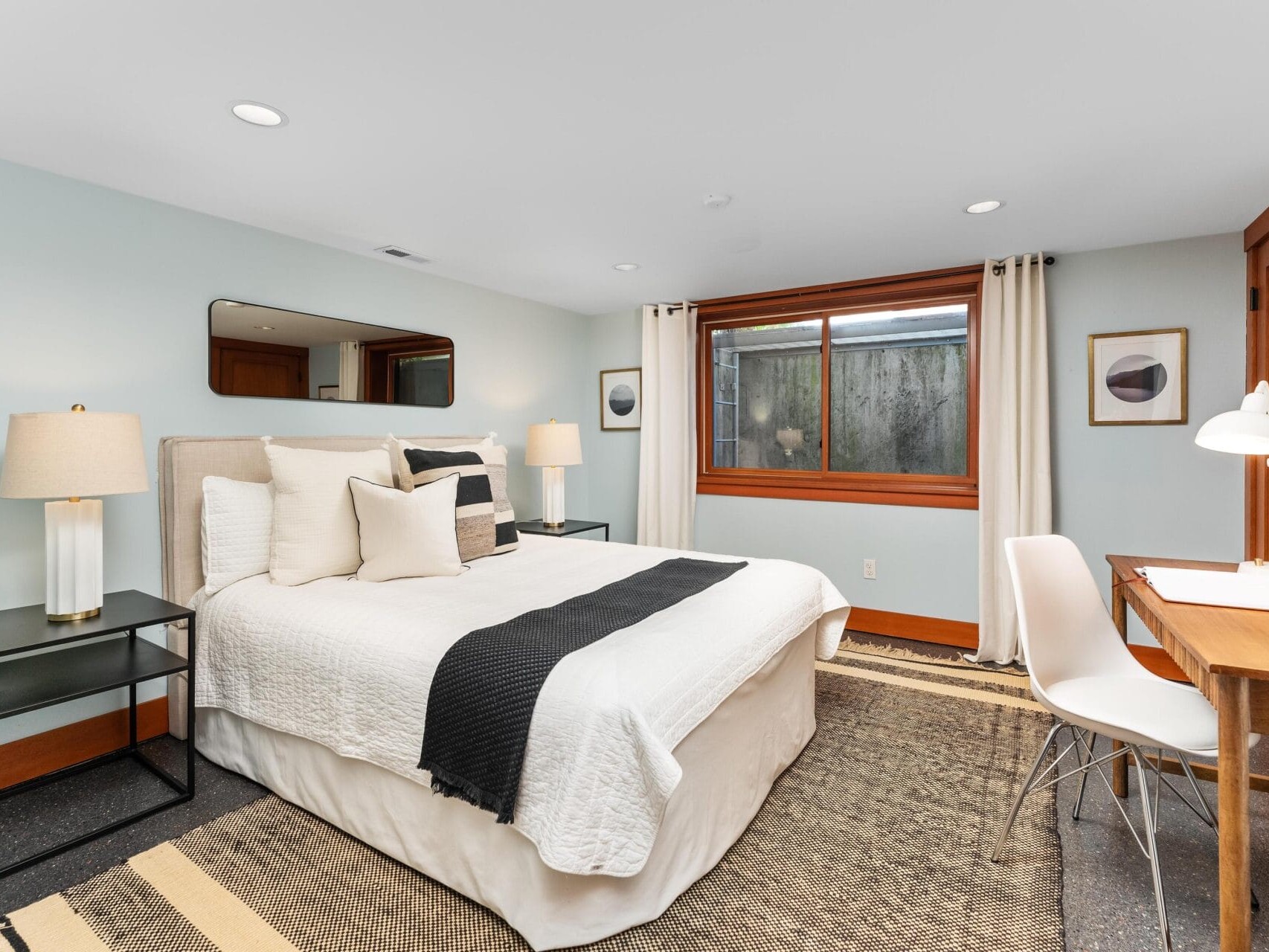 A cozy Portland, Oregon bedroom with a queen-sized bed, white bedding, and black throw pillows. Wooden nightstands with lamps flank the bed, and a large window with curtains invites natural light. A wooden desk with a white chair complements the minimalist decor.