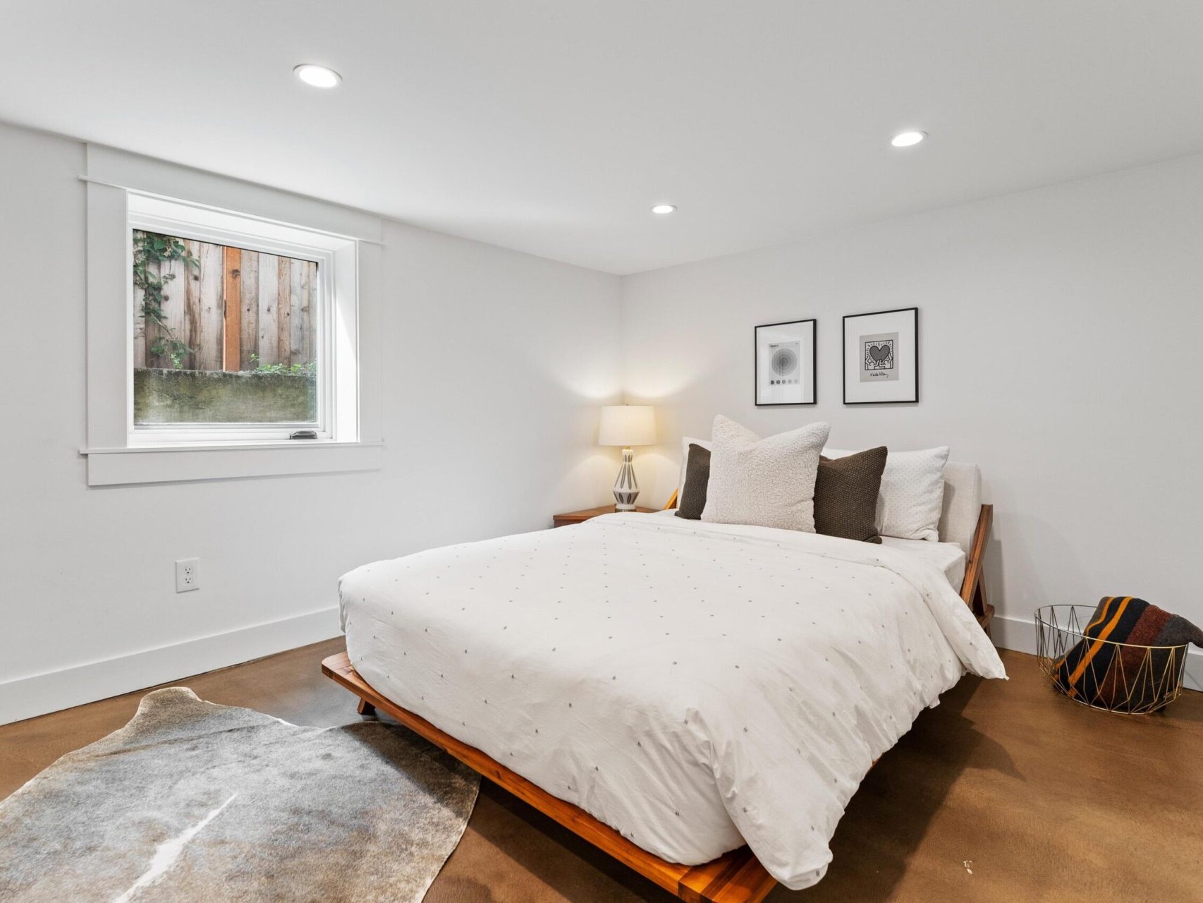 A minimalist bedroom in Portland, Oregon features a bed with white bedding and several pillows, a small nightstand with a lamp, and two framed pictures on the wall. A cowhide rug graces the brown floor, while a window offers natural light.