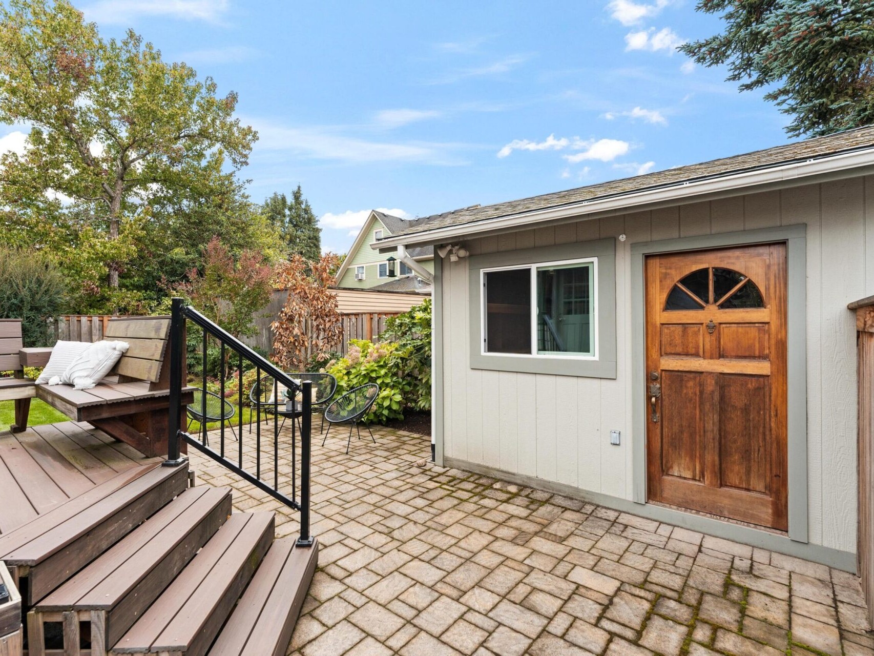 A small gray shed with a wooden door is in a backyard with a paved patio. There are steps leading to a wooden deck with bench seating. Trees and bushes are visible in the background under a partly cloudy sky.