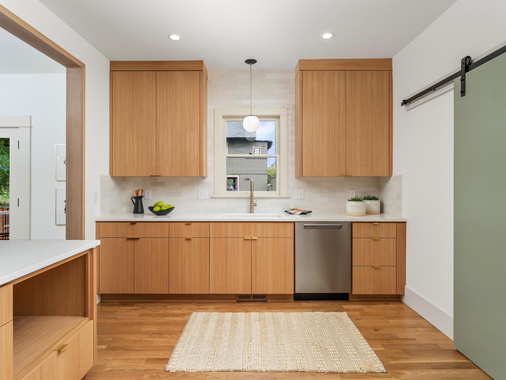 A modern kitchen in Portland, Oregon, features wooden cabinets and flooring. A window above the sink lets in natural light. There’s a hanging pendant light, a dishwasher, and a light green sliding door on the right. A woven rug lies gracefully on the floor.